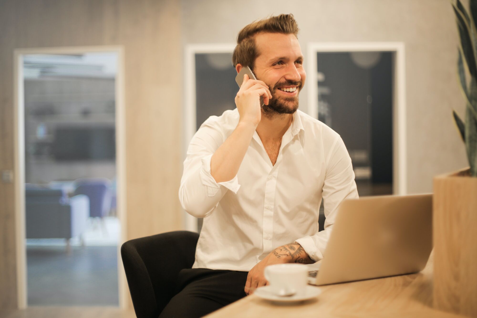 CEKOME AGENCE WEB02 | Cekome Homme d'affaires souriant au téléphone dans un bureau moderne avec un ordinateur portable et une tasse de café.
