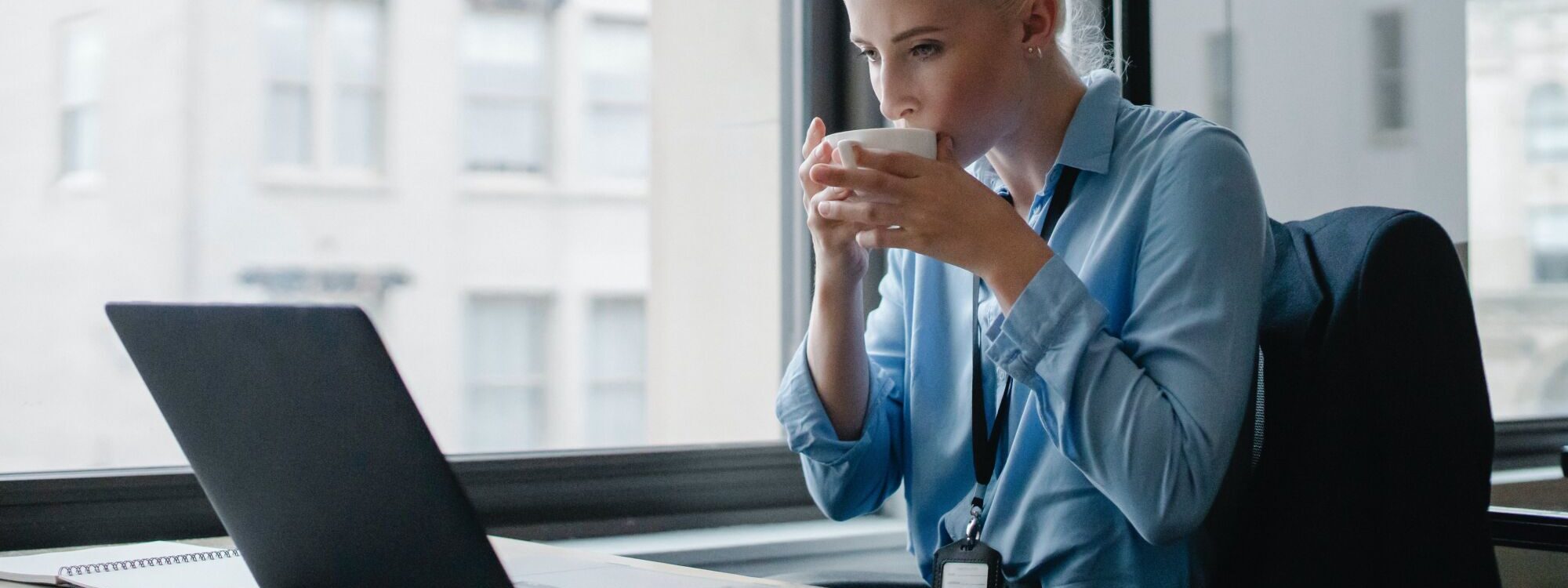 Femme d'affaires buvant une tasse de caf&eacute; tout en travaillant sur un ordinateur portable devant une fen&ecirc;tre.