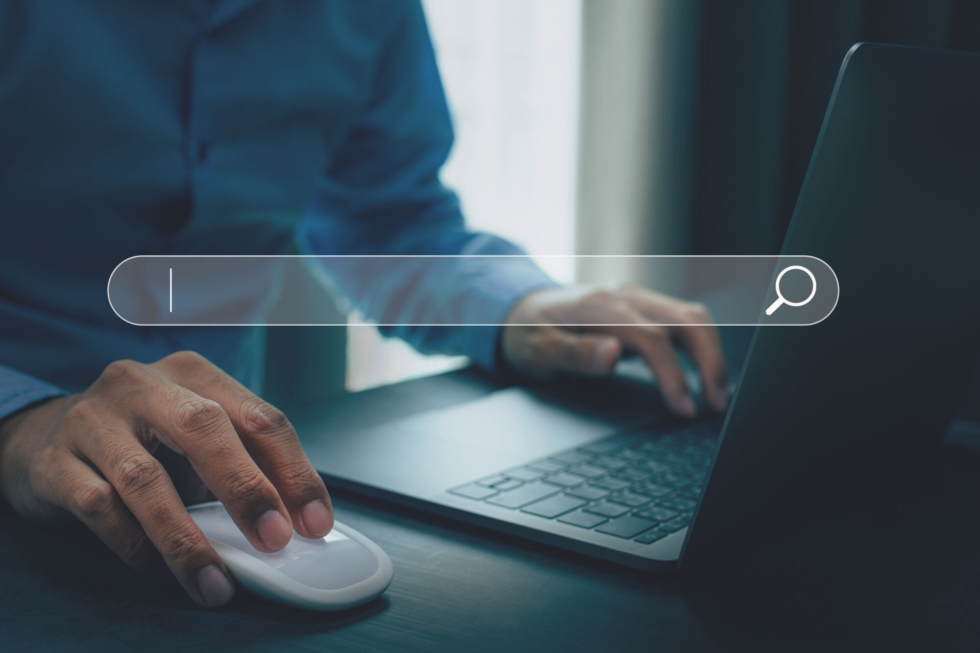 ADOBESTOCK_476219144 | Cekome Main utilisant une souris d'ordinateur avec une barre de recherche sur un écran d'ordinateur portable dans une pièce sombre.