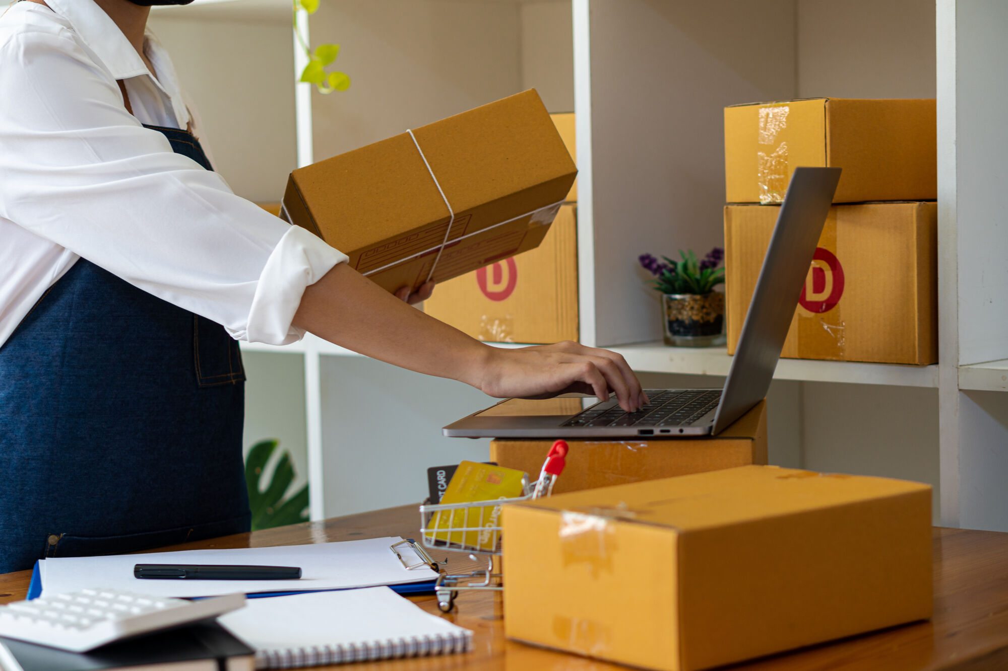 ADOBESTOCK_486506969 | Cekome Personne emballant des colis tout en utilisant un ordinateur portable pour les opérations de vente en ligne.