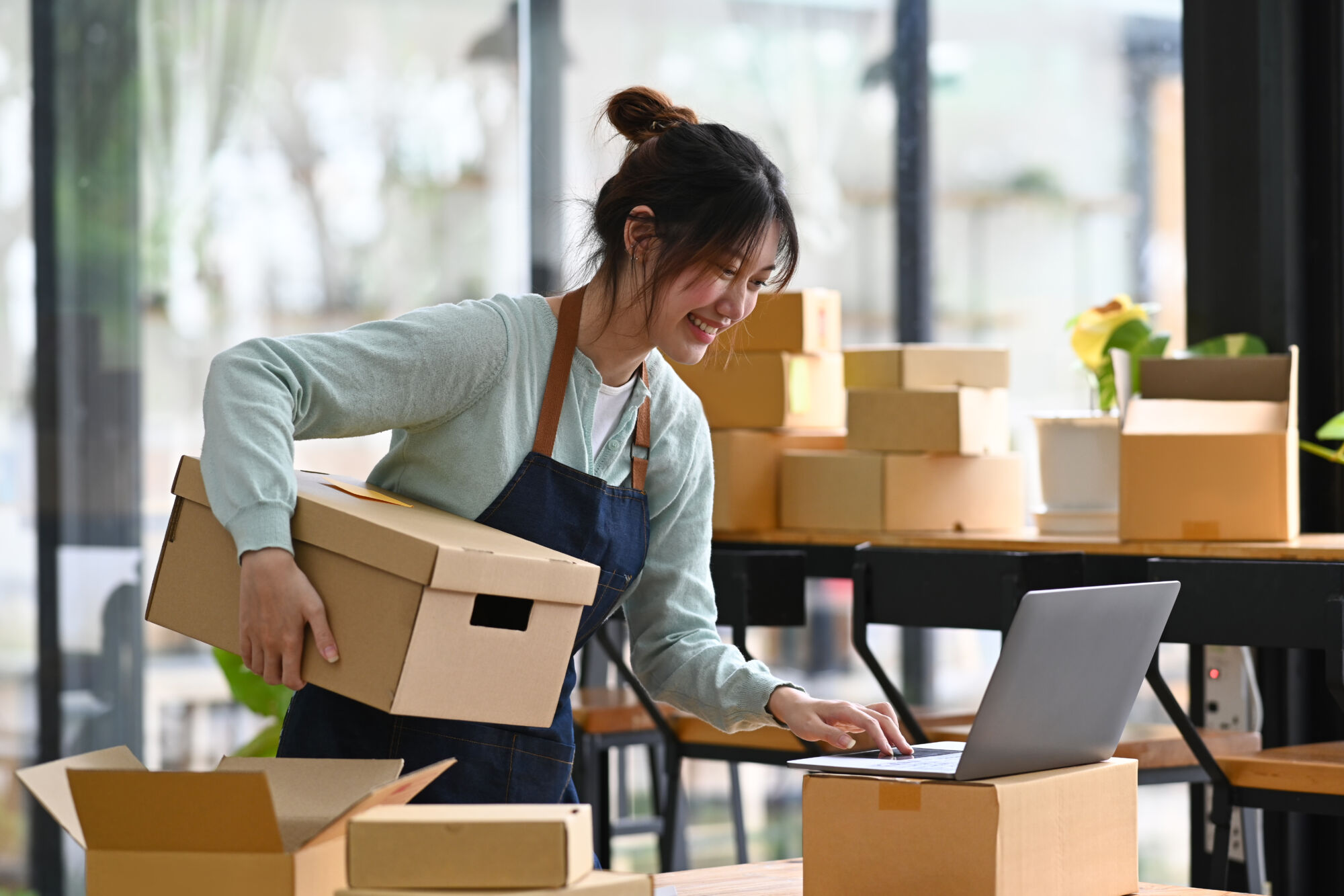 ADOBESTOCK_507437543 | Cekome Femme souriante préparant des commandes tout en interagissant avec un ordinateur portable dans un espace de travail lumineux.