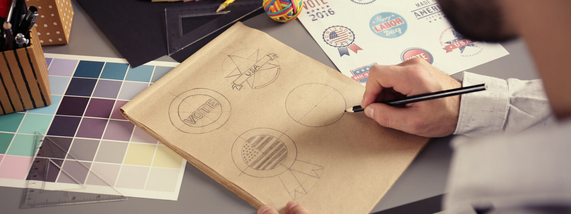 ADOBESTOCK_136869512 2000X750 | Cekome Graphiste dessinant des logos sur un cahier avec des échantillons de couleur et des autocollants électoraux.