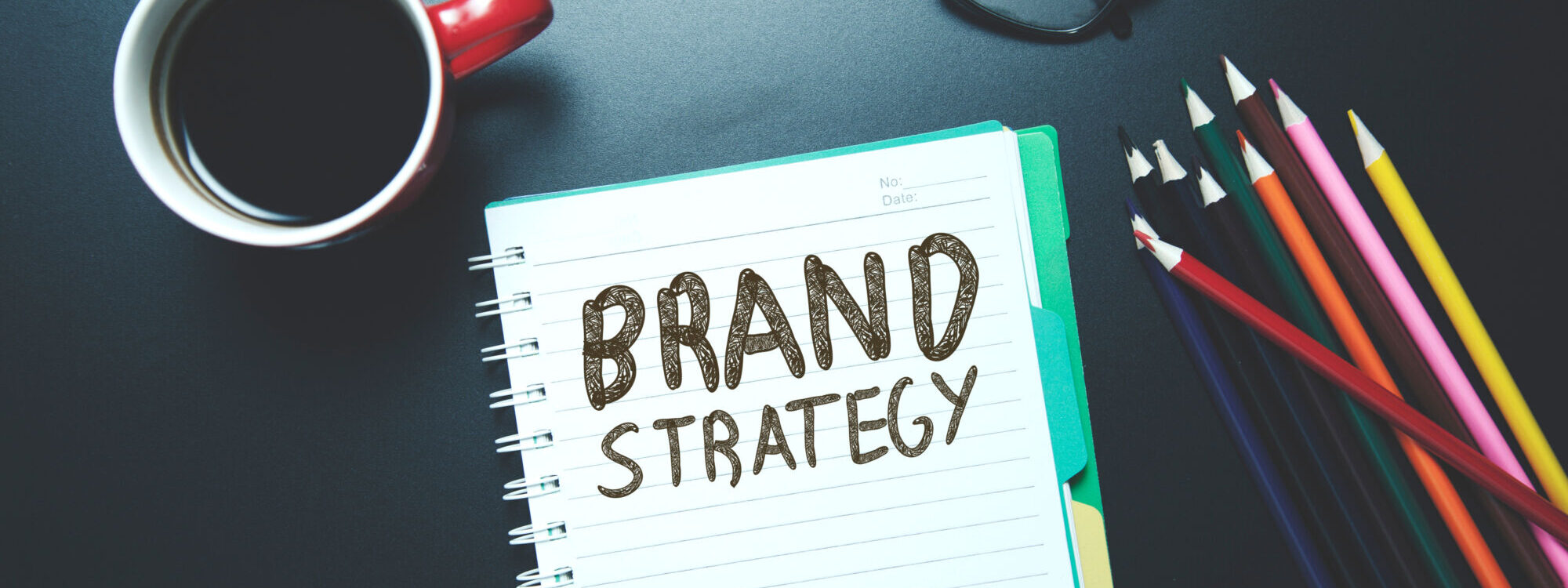 ADOBESTOCK_192898766 2000X750 | Cekome Carnet avec l'inscription 'BRAND STRATEGY' accompagné d'une tasse de café et de lunettes sur un bureau sombre.