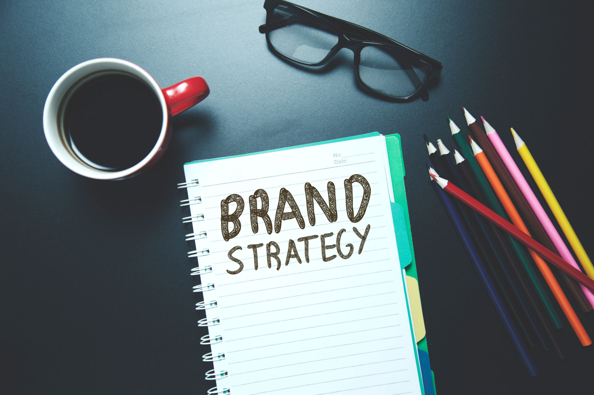 ADOBESTOCK_192898766 | Cekome Carnet avec l'inscription 'BRAND STRATEGY' accompagné d'une tasse de café et de lunettes sur un bureau sombre.
