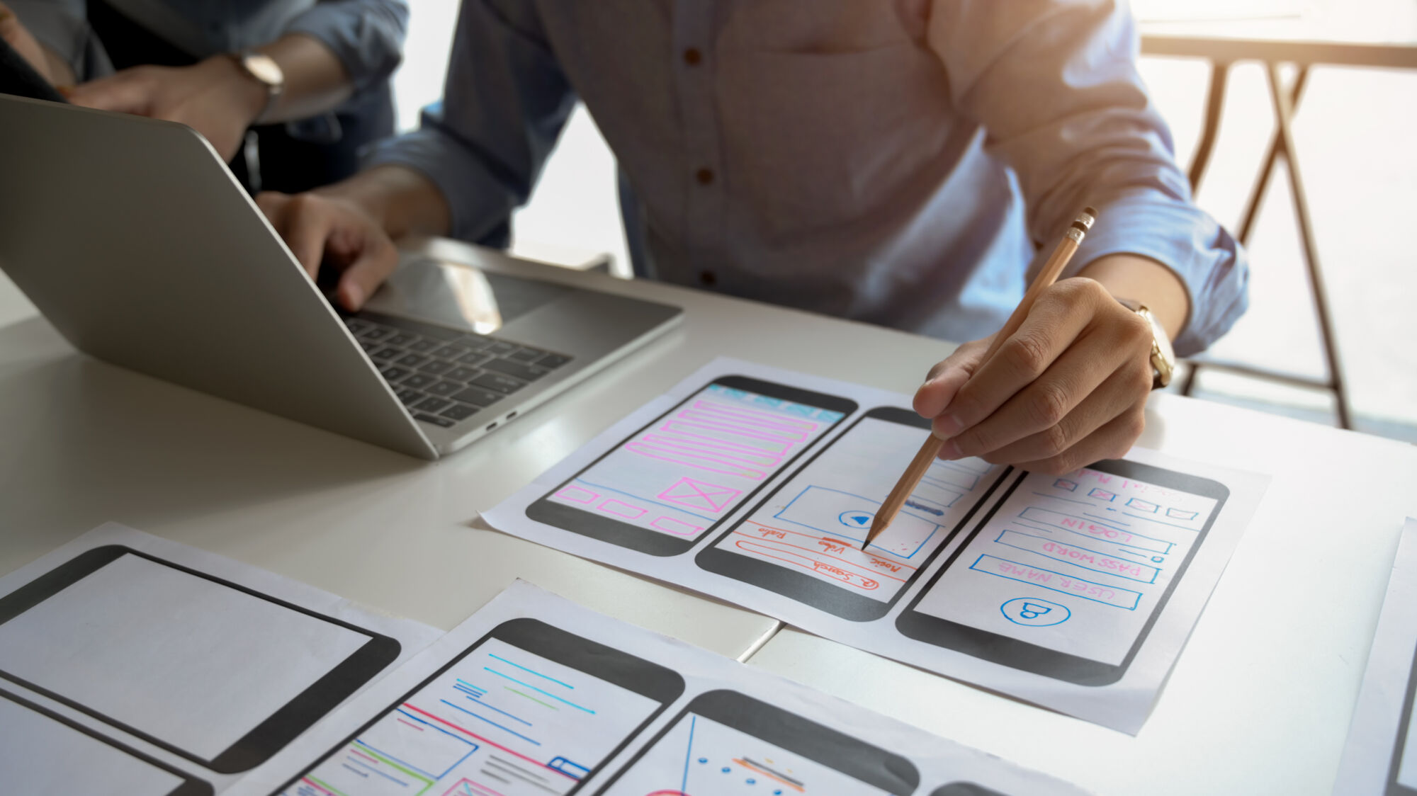 ADOBESTOCK_322689260 | Cekome Personnes analysant des ébauches de conception d'interface mobile avec un ordinateur portable en arrière-plan.