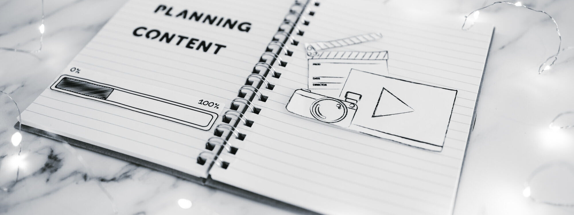 Cahier ouvert avec 'PLANNING CONTENT' &eacute;crit et dessins de mat&eacute;riel multim&eacute;dia sur un bureau en marbre.