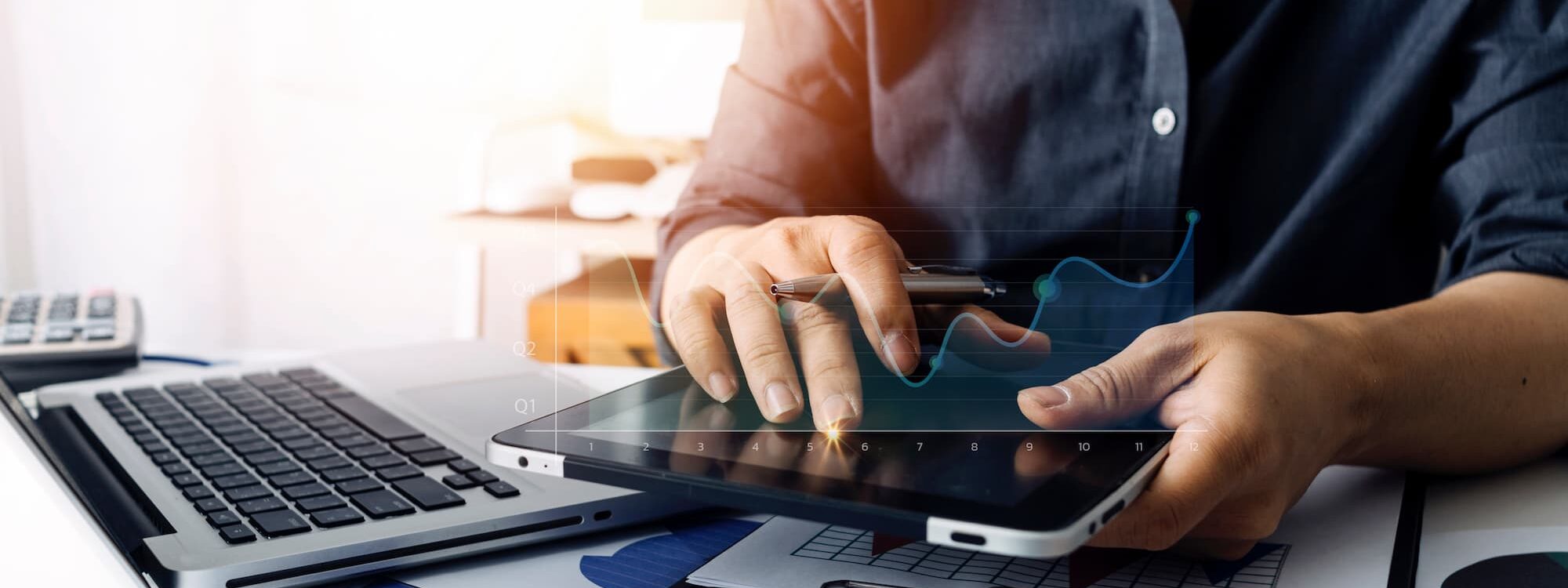 Personne analysant des statistiques financi&egrave;res sur une tablette num&eacute;rique avec ordinateur portable et documents en arri&egrave;re-plan.