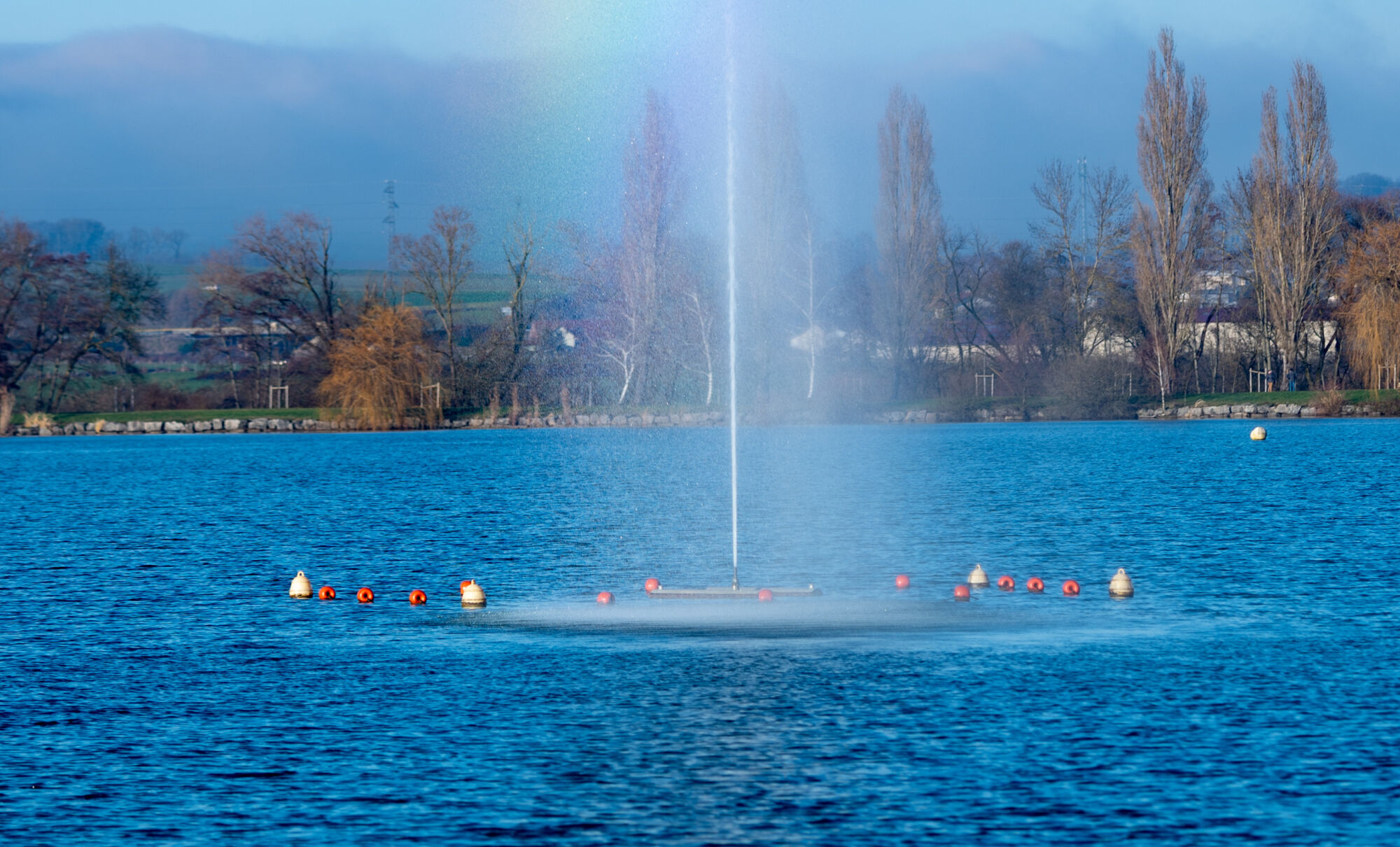 Jet d'eau dans un lac avec arbres et montagnes en arri&egrave;re-plan sous un ciel bleu.