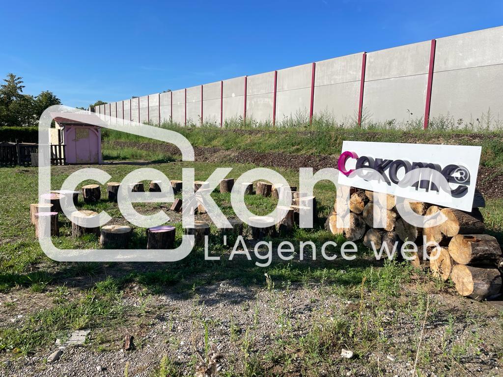 8 30 | Cekome Panneau de Cekome avec des bûches en avant-plan sur fond d'herbe et de bâtiment industriel