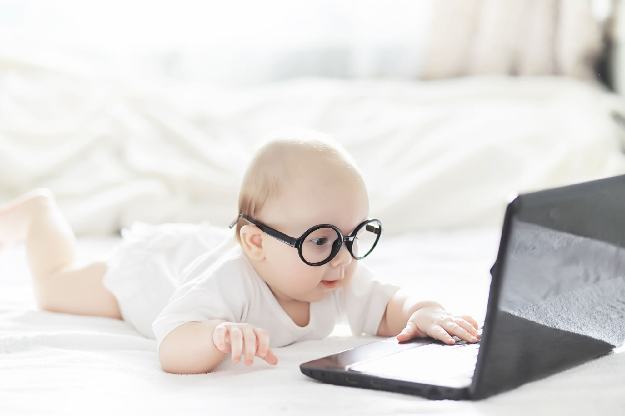 ADOBESTOCK_306643510 | Cekome Bébé avec des lunettes regardant un ordinateur portable ouvert sur un lit, concept d'apprentissage précoce ou de technologie.