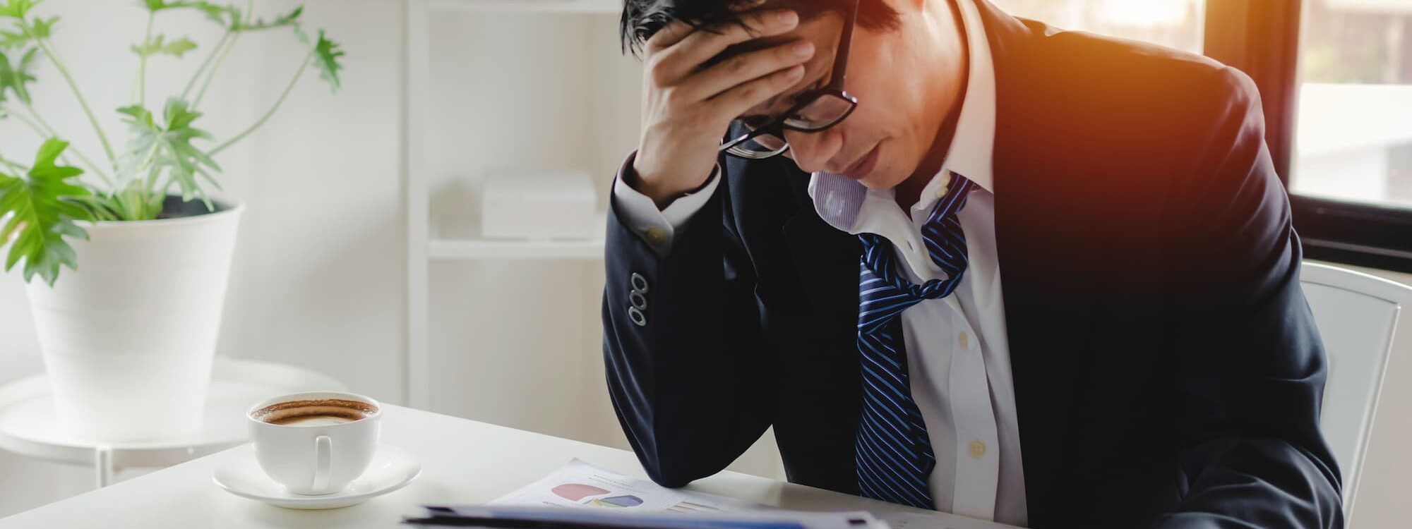 Homme d'affaires frustré tenant sa tête au-dessus d'un rapport financier dans un bureau lumineux avec une tasse de café et une calculatrice
