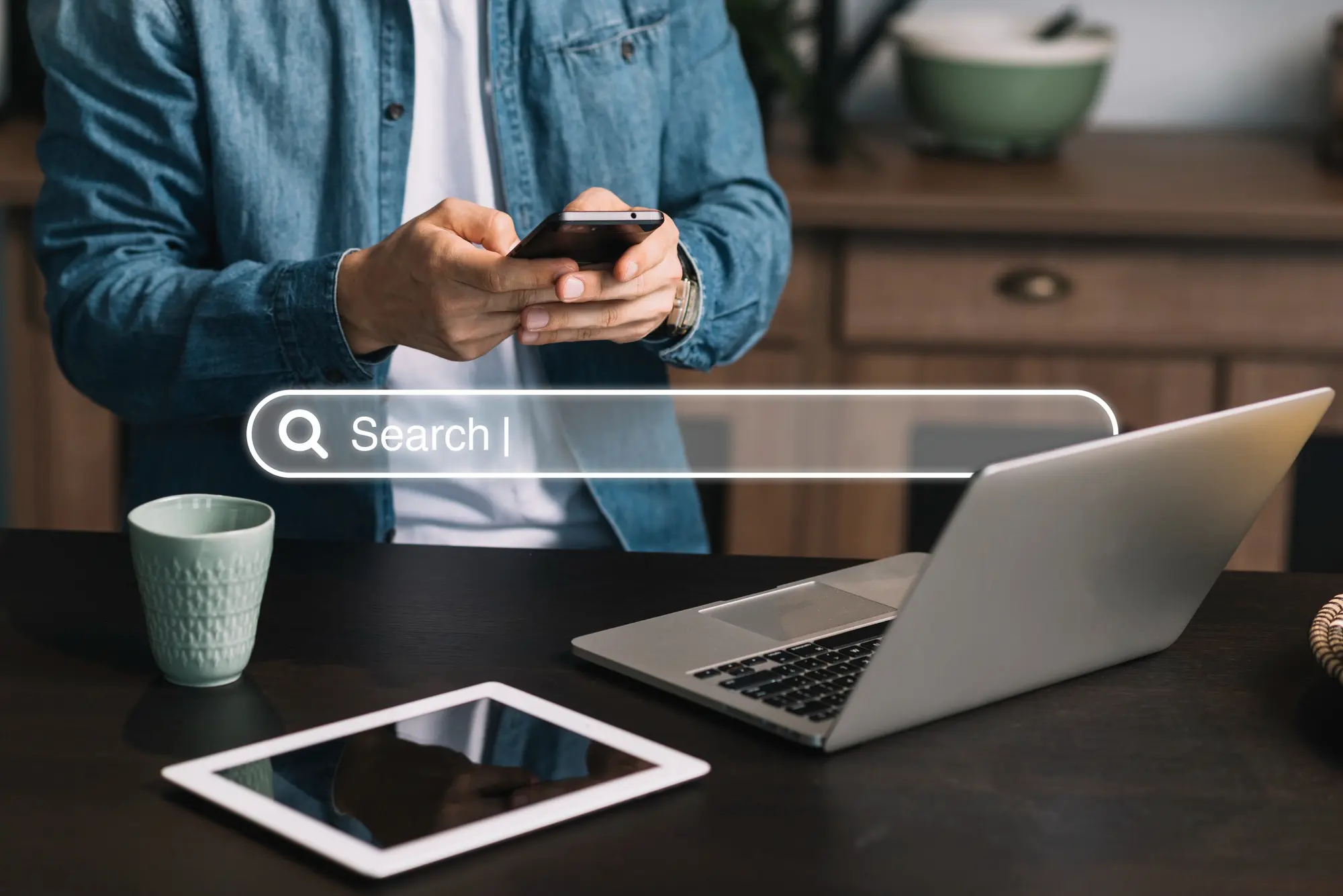 Personne utilisant un smartphone devant un ordinateur portable avec une barre de recherche virtuelle