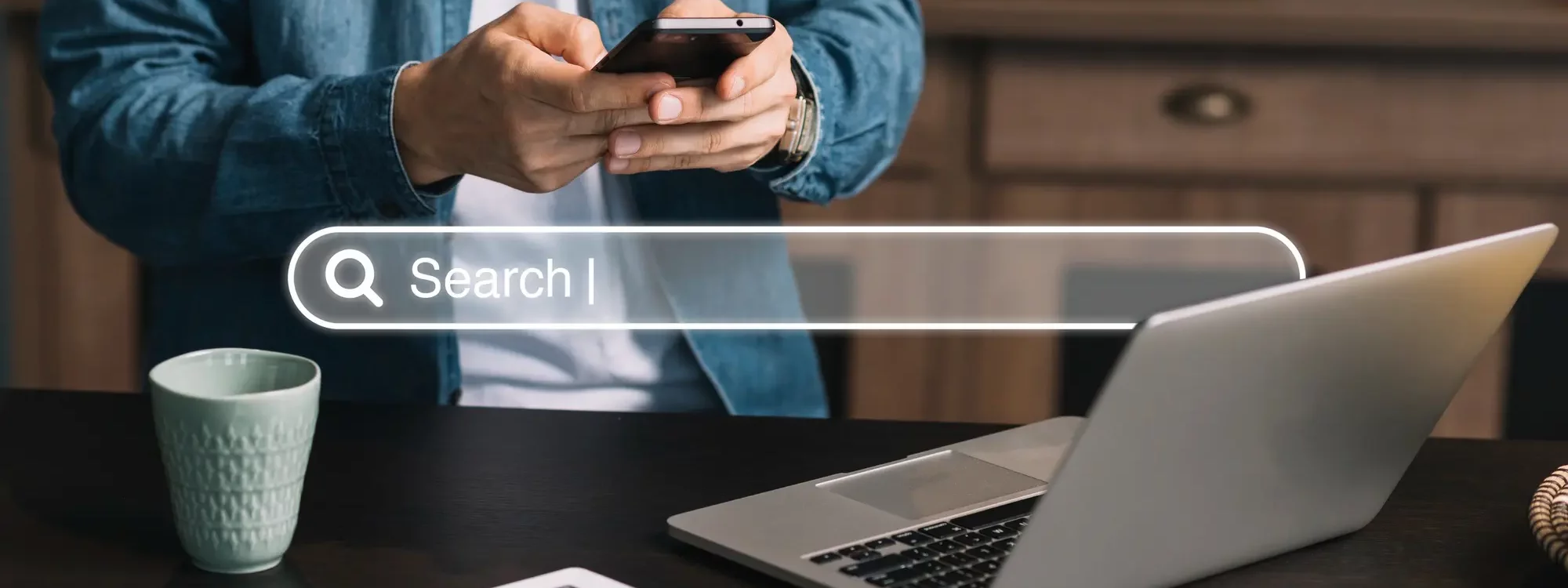 Personne utilisant un smartphone devant un ordinateur portable avec une barre de recherche virtuelle