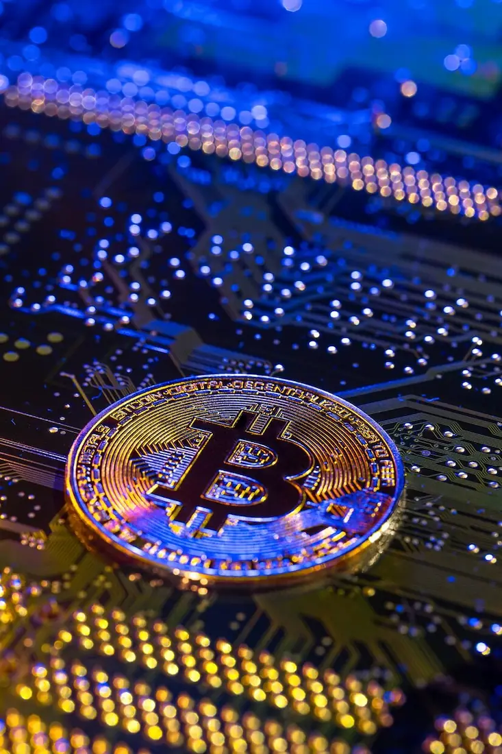 BITCOIN COIN CLOSEUP CIRCUIT BOARD FINANCE CONCEPT_169016 61885 | Cekome Pièce Bitcoin dorée posée sur un circuit imprimé illuminé par des lumières bleues et jaunes.