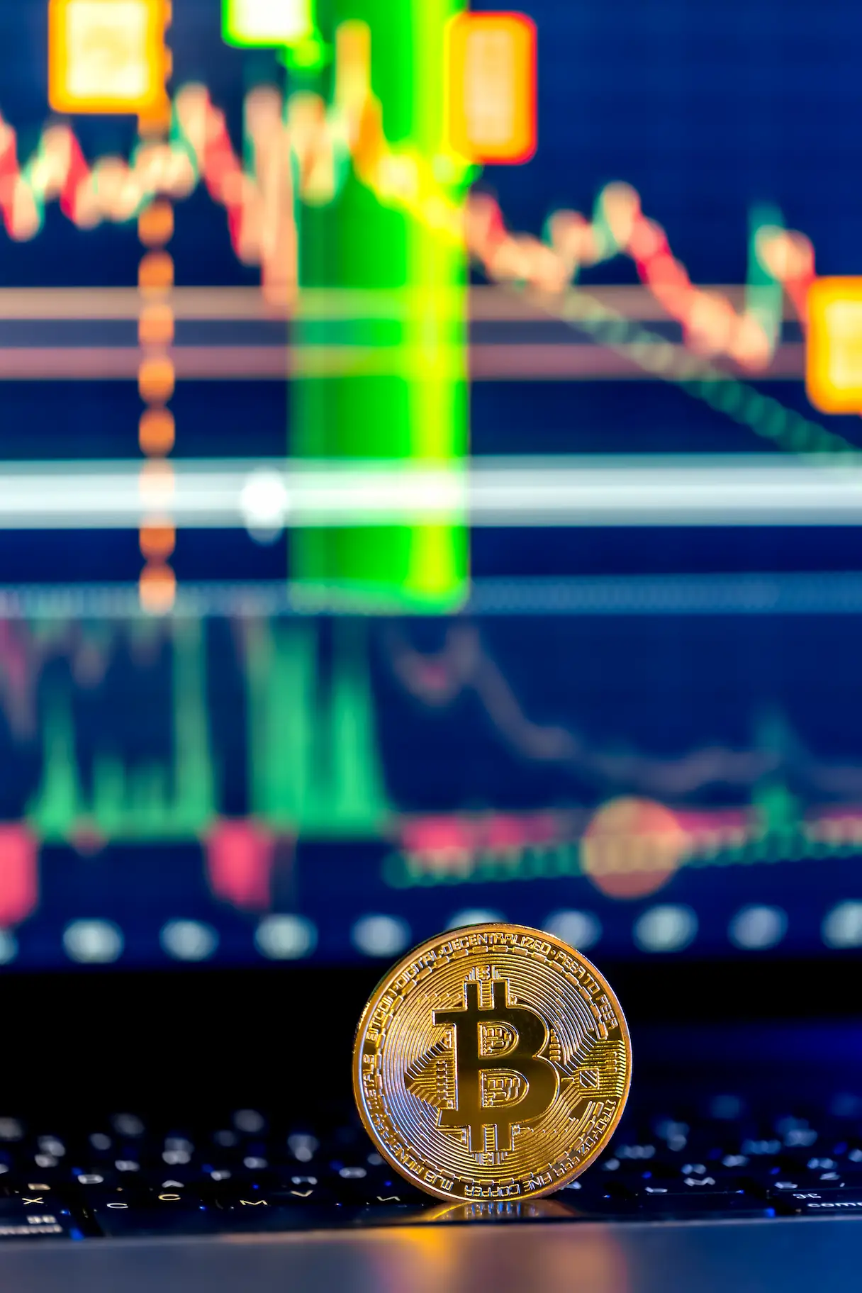 Pièce de bitcoin dorée posée sur un clavier d'ordinateur, avec un graphique boursier flou en arrière-plan.