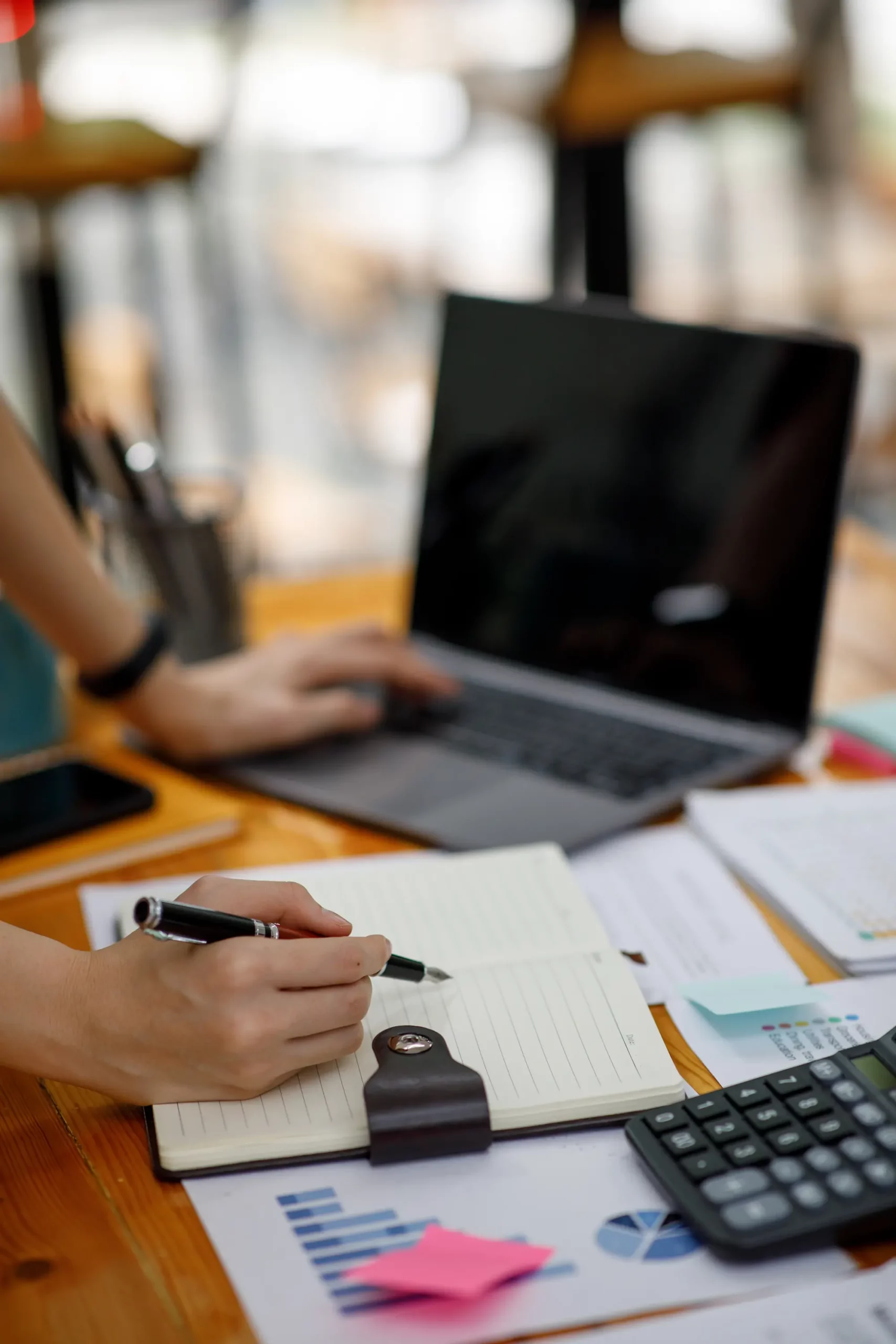 Personne prenant des notes devant un ordinateur portable avec des graphiques et une calculatrice sur le bureau