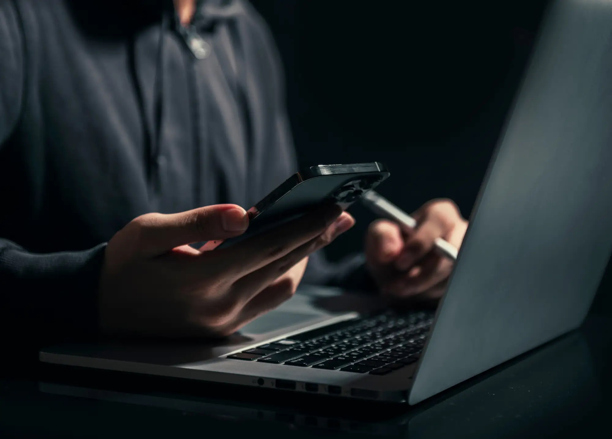 ADOBESTOCK_451638578 | Cekome Personne en sweat à capuche utilisant un smartphone et un ordinateur portable dans un environnement sombre, symbolisant la cybersécurité ou le piratage.