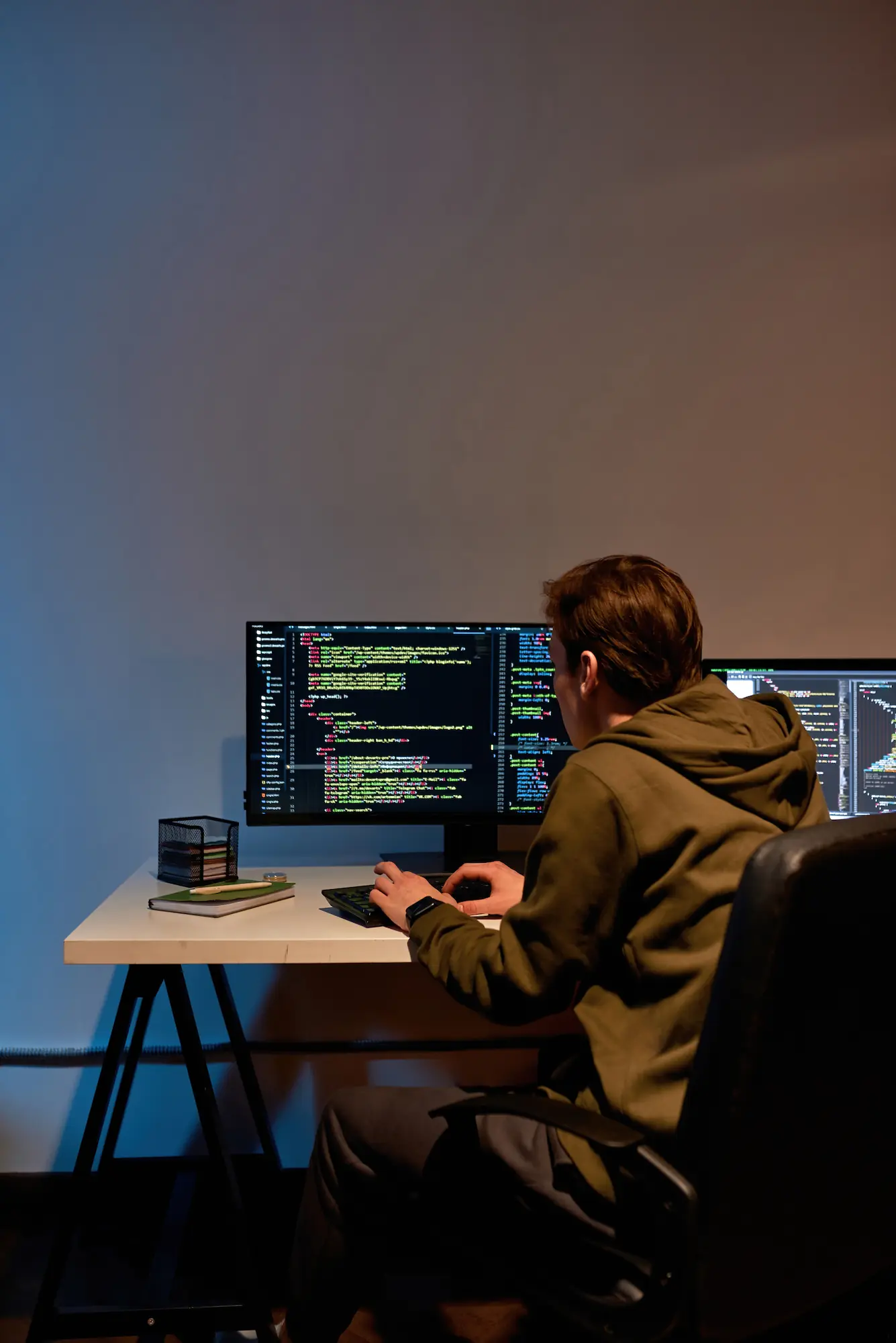 ADOBESTOCK_487777745 | Cekome Développeur en hoodie vert travaillant sur du code informatique affiché sur deux écrans dans un bureau sombre.
