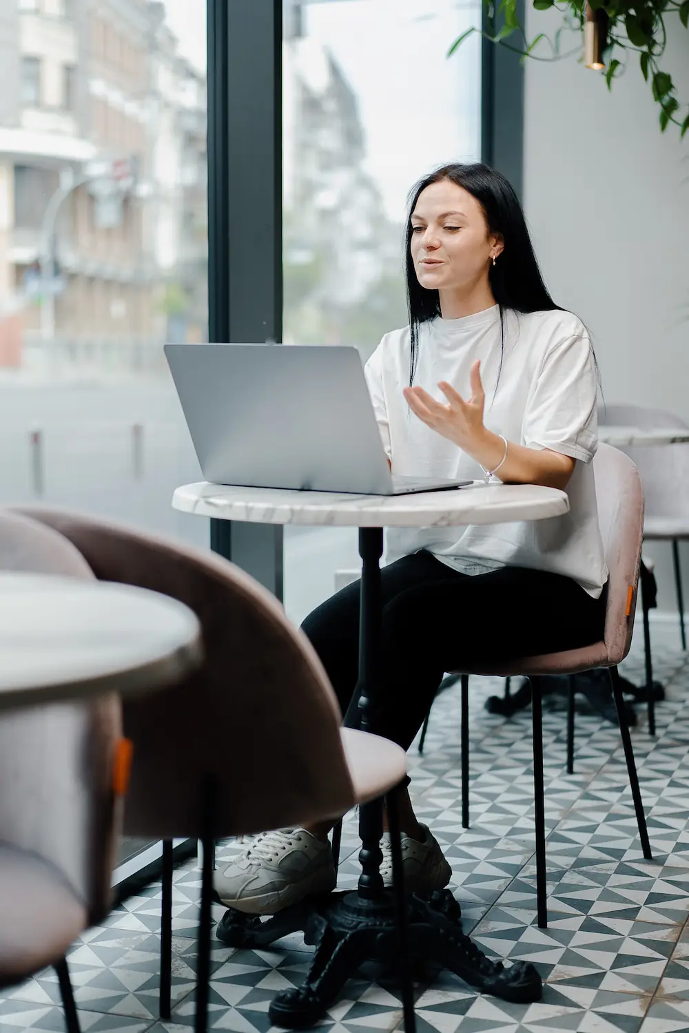ADOBESTOCK_512542862 | Cekome Femme souriante en discussion vidéo sur un ordinateur portable dans un café moderne, près d'une grande fenêtre donnant sur la rue.