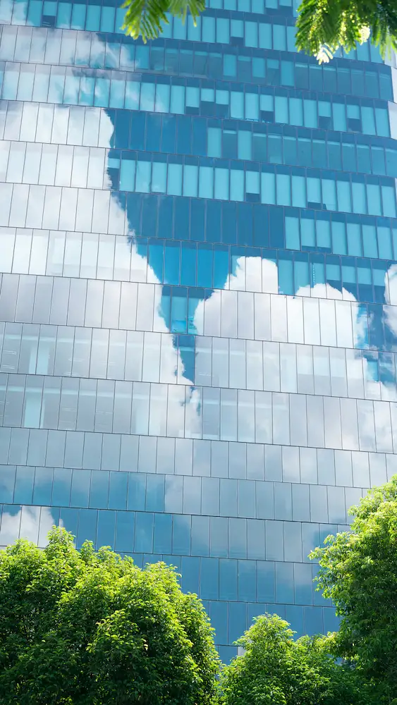 ADOBESTOCK_527028004 | Cekome Façade en verre d'un immeuble reflétant le ciel et les nuages, entourée de verdure.