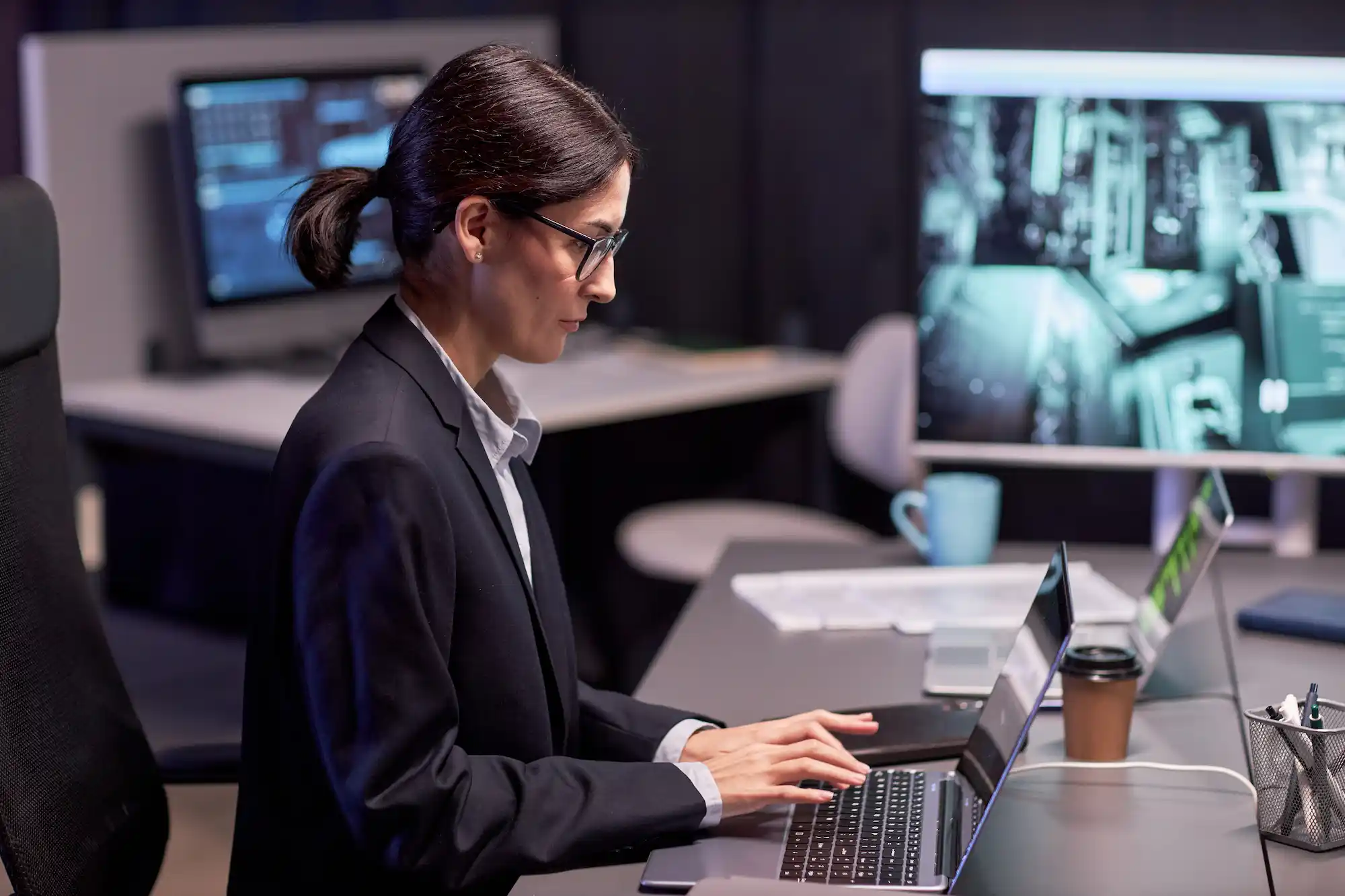 ADOBESTOCK_584005525 | Cekome Femme en costume travaillant sur un ordinateur portable dans un centre de cybersécurité, avec des écrans affichant des données et des caméras de surveillance.