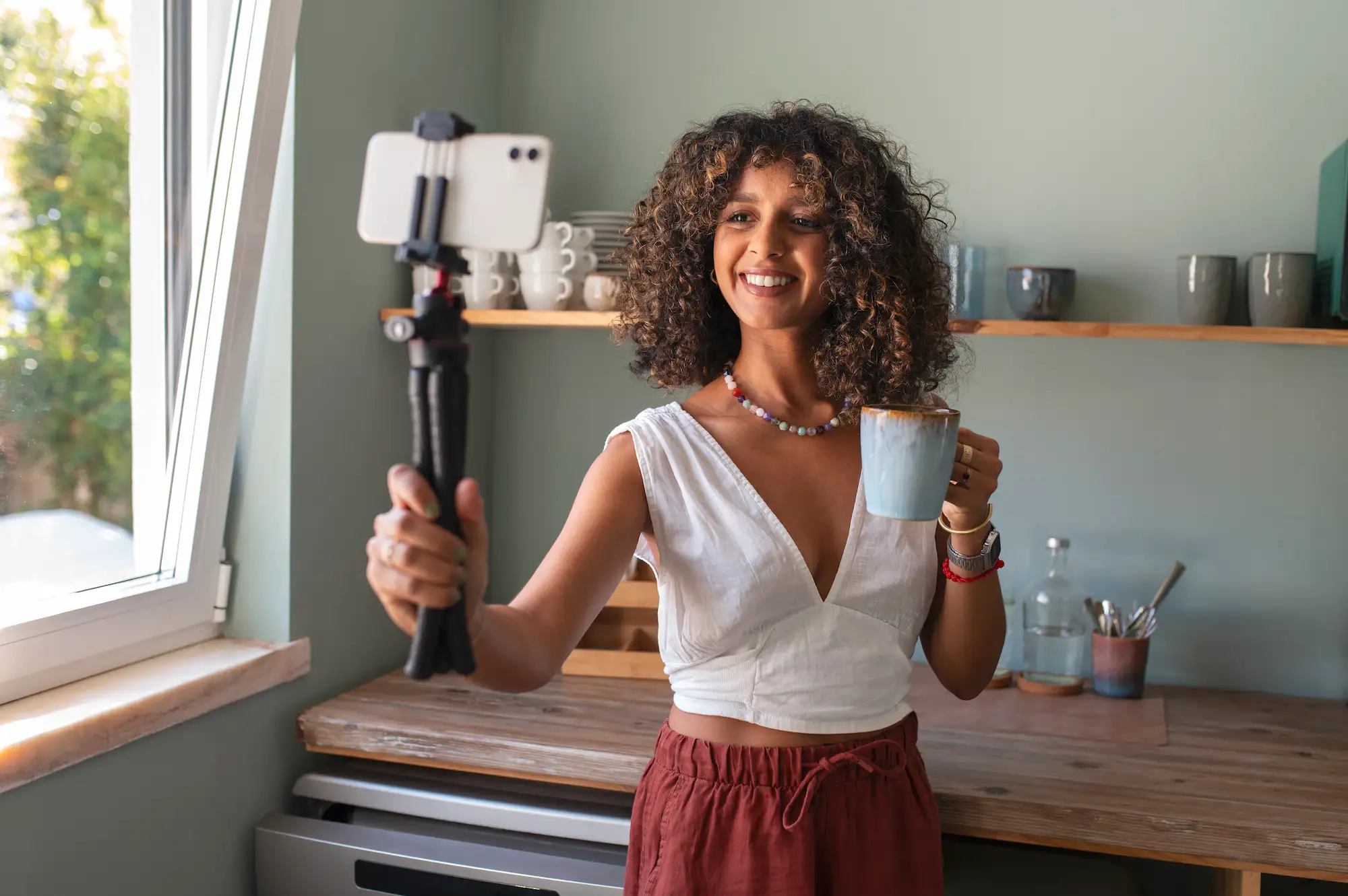 Jeune femme souriante enregistrant une vidéo avec son smartphone sur trépied tout en tenant une tasse de café dans une cuisine lumineuse.