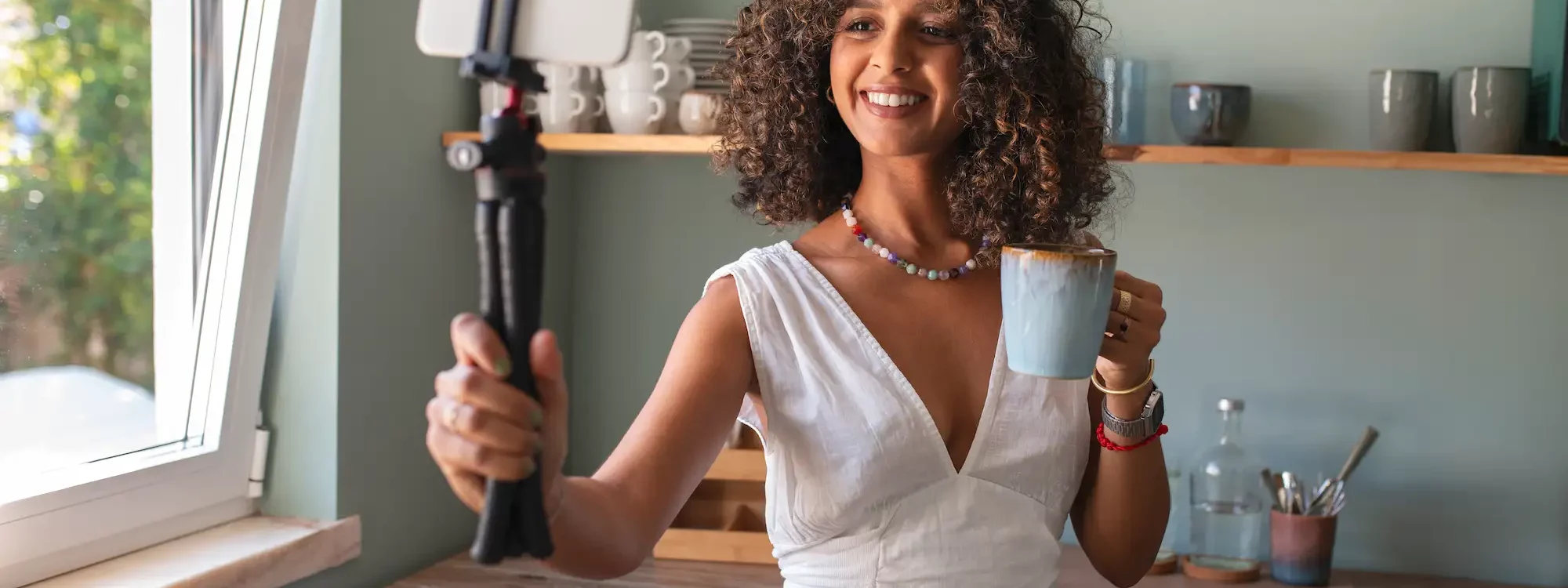 Jeune femme souriante enregistrant une vidéo avec son smartphone sur trépied tout en tenant une tasse de café dans une cuisine lumineuse.