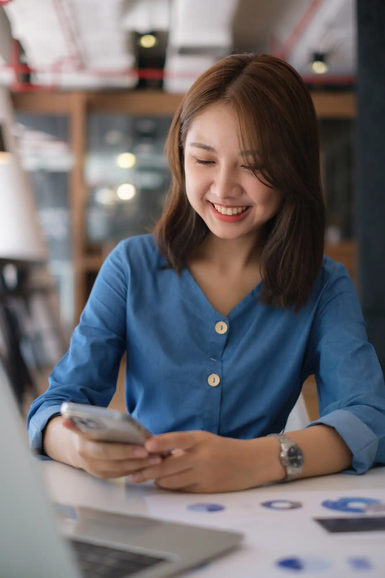 ADOBESTOCK_496039260 | Cekome Femme souriante en chemise bleue utilisant un smartphone à son bureau.