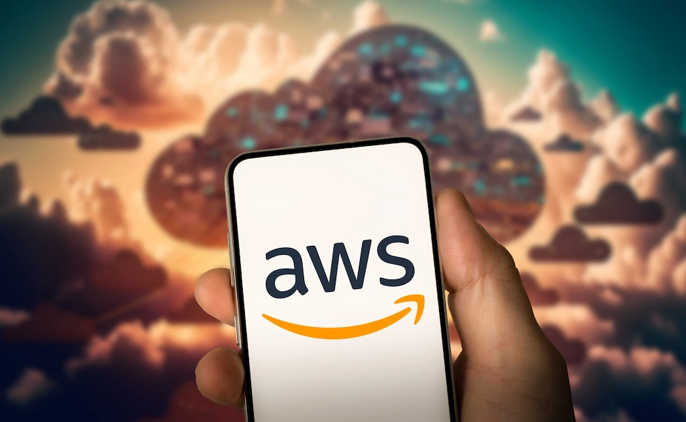 ADOBESTOCK_683774552_EDITORIAL_USE_ONLY 1 | Cekome Main tenant un smartphone affichant le logo AWS devant un arrière-plan de nuages numériques