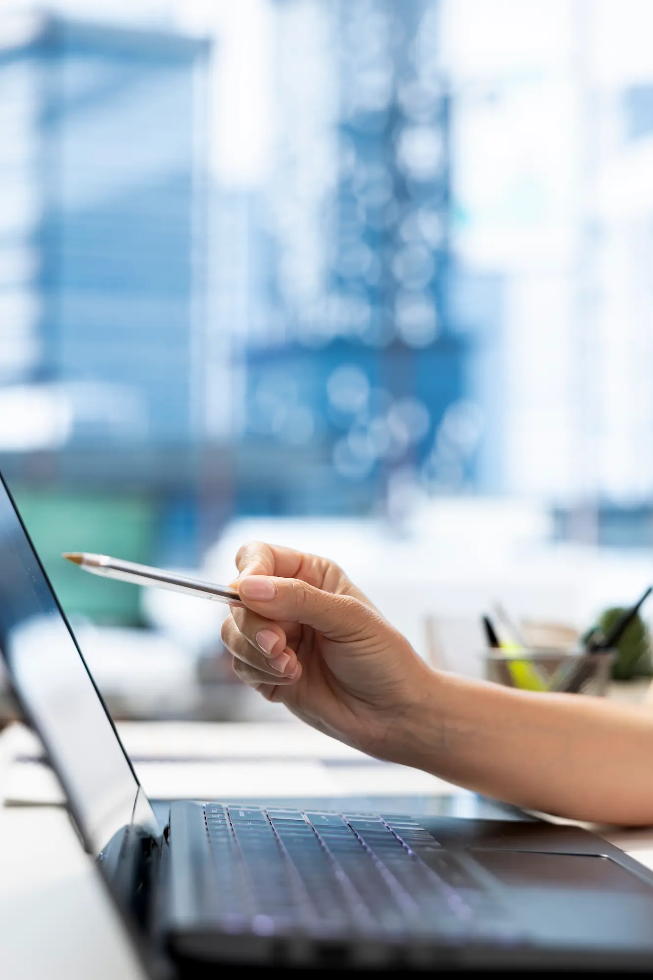 HR DEPARTMENT WORKER USING LAPTOP BACKGROUND CHECKS JOB APPLICANTS | Cekome Main tenant un stylo pointé vers l’écran d’un ordinateur portable dans un bureau moderne avec vue sur des immeubles flous en arrière-plan