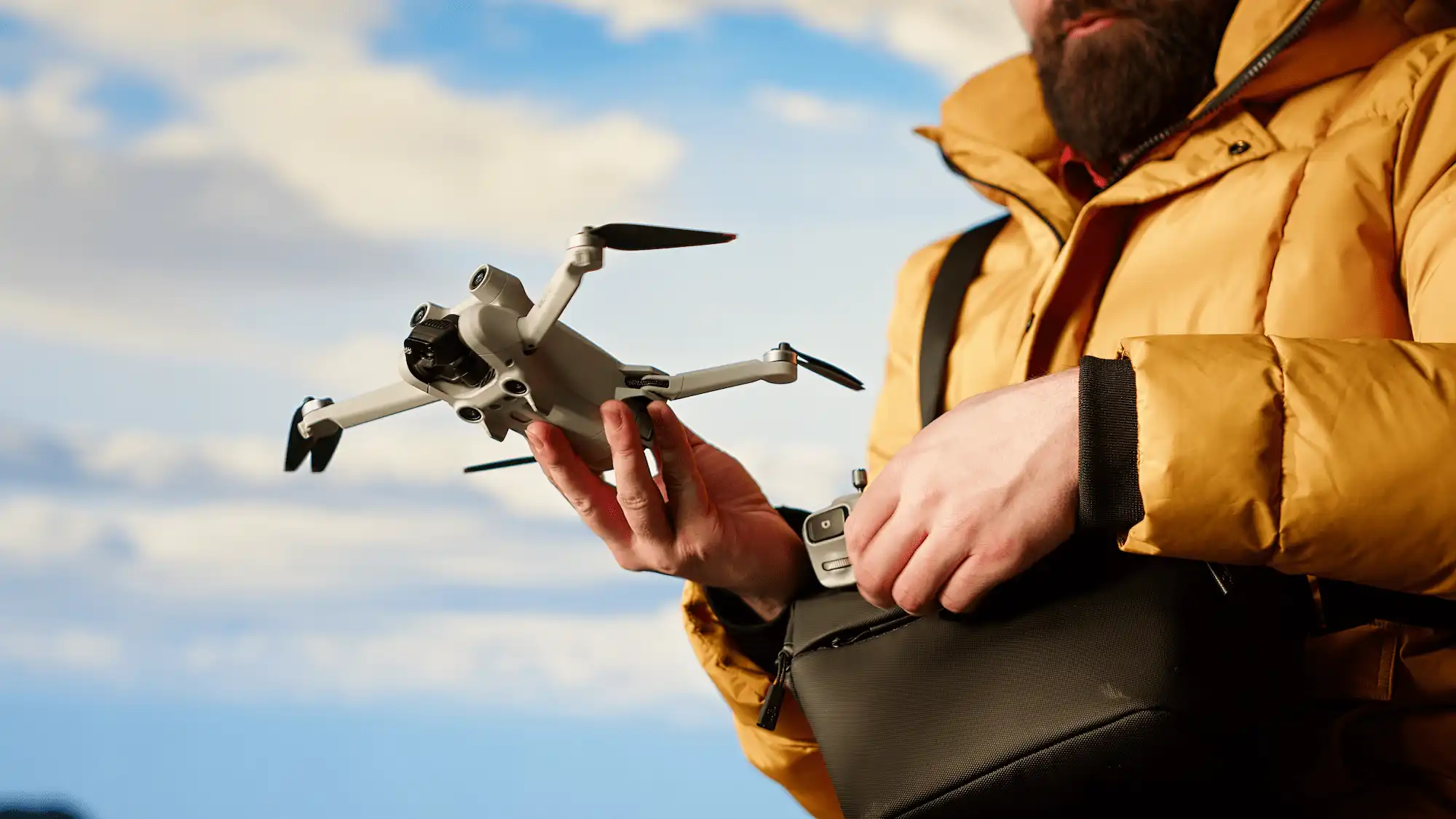 MODERN TRAVELER CREATING CONTENT FILMING VALLEY WITH HIS DRONE | Cekome Homme barbu en manteau jaune préparant un drone pour filmer en extérieur, illustrant l’usage moderne des agents autonomes pour la création de contenu