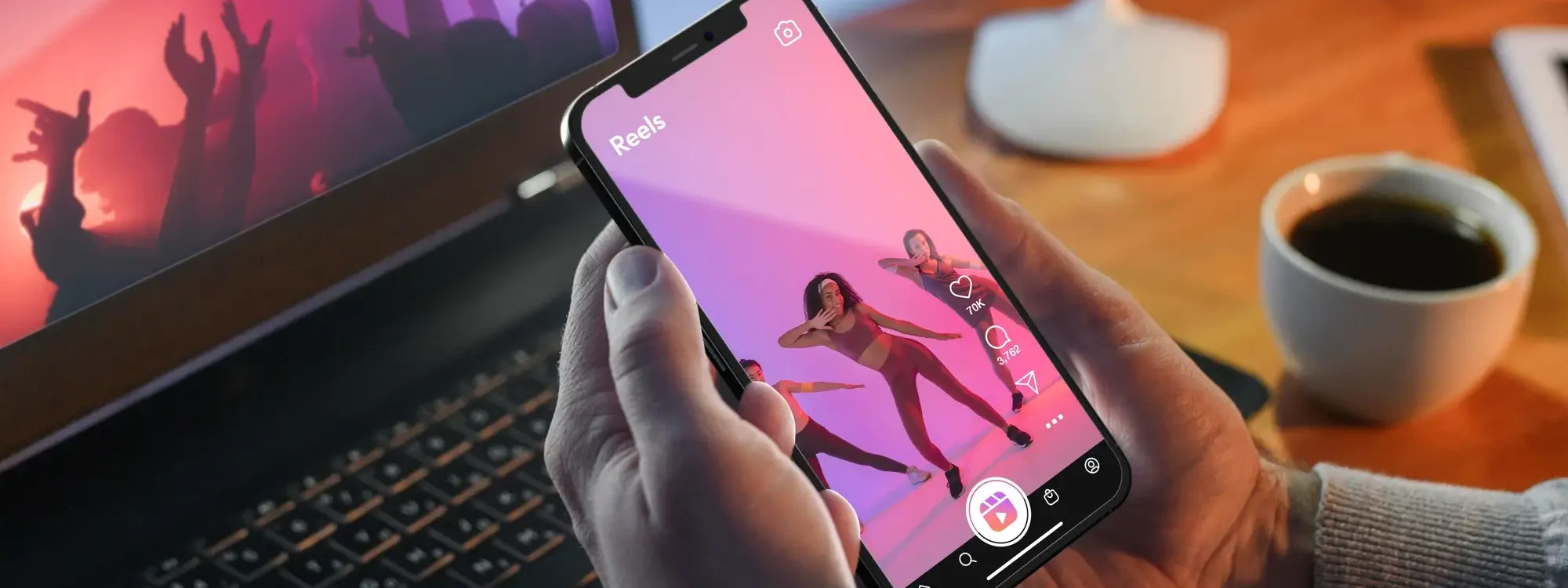 Personne tenant un smartphone affichant une vid&eacute;o Reels de danse, devant un ordinateur portable et une tasse de caf&eacute; sur un bureau en bois