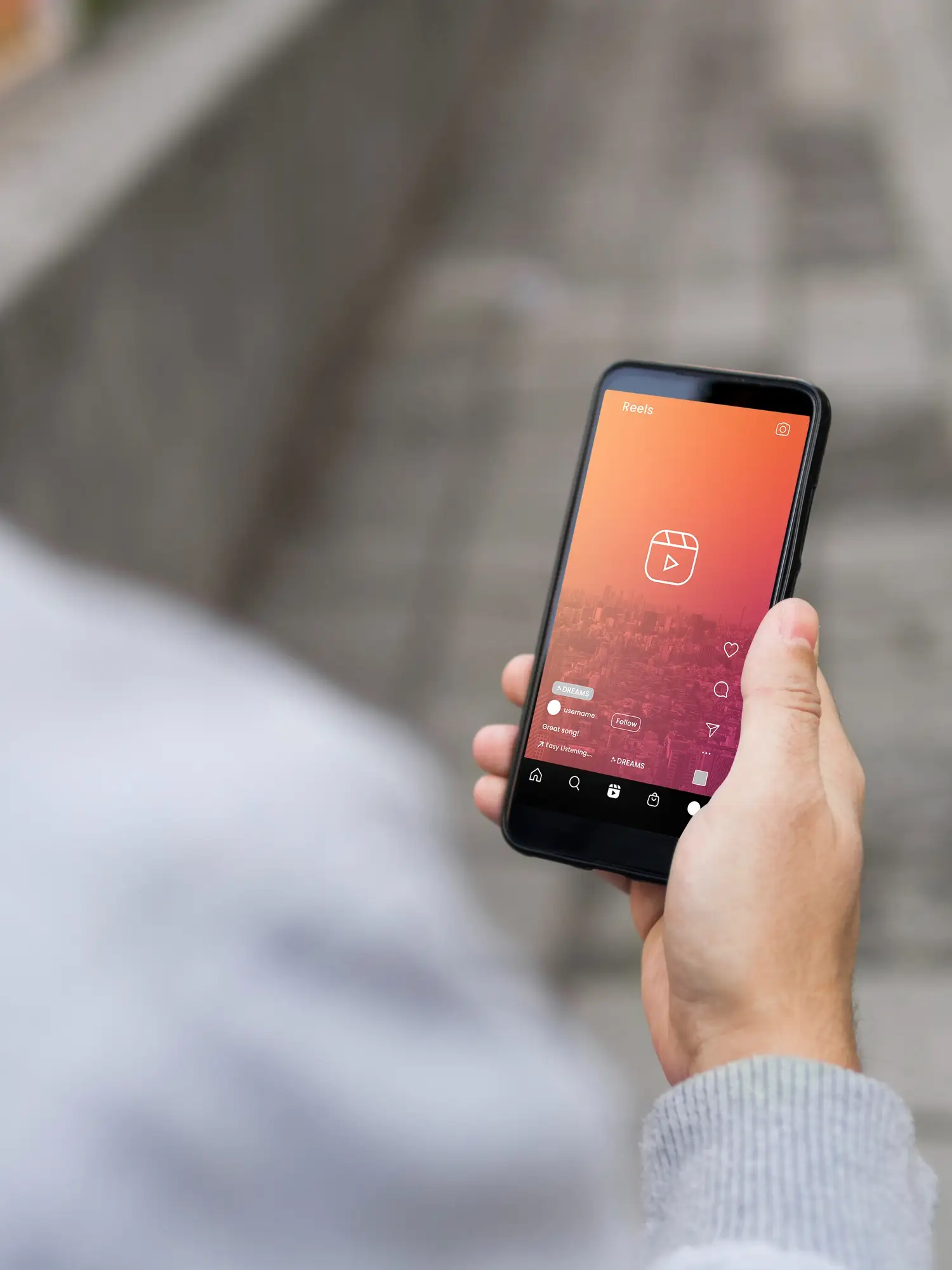 Personne tenant un smartphone affichant un &eacute;cran d&rsquo;accueil Reels d&rsquo;Instagram en ext&eacute;rieur, sur fond de rue pav&eacute;e floue