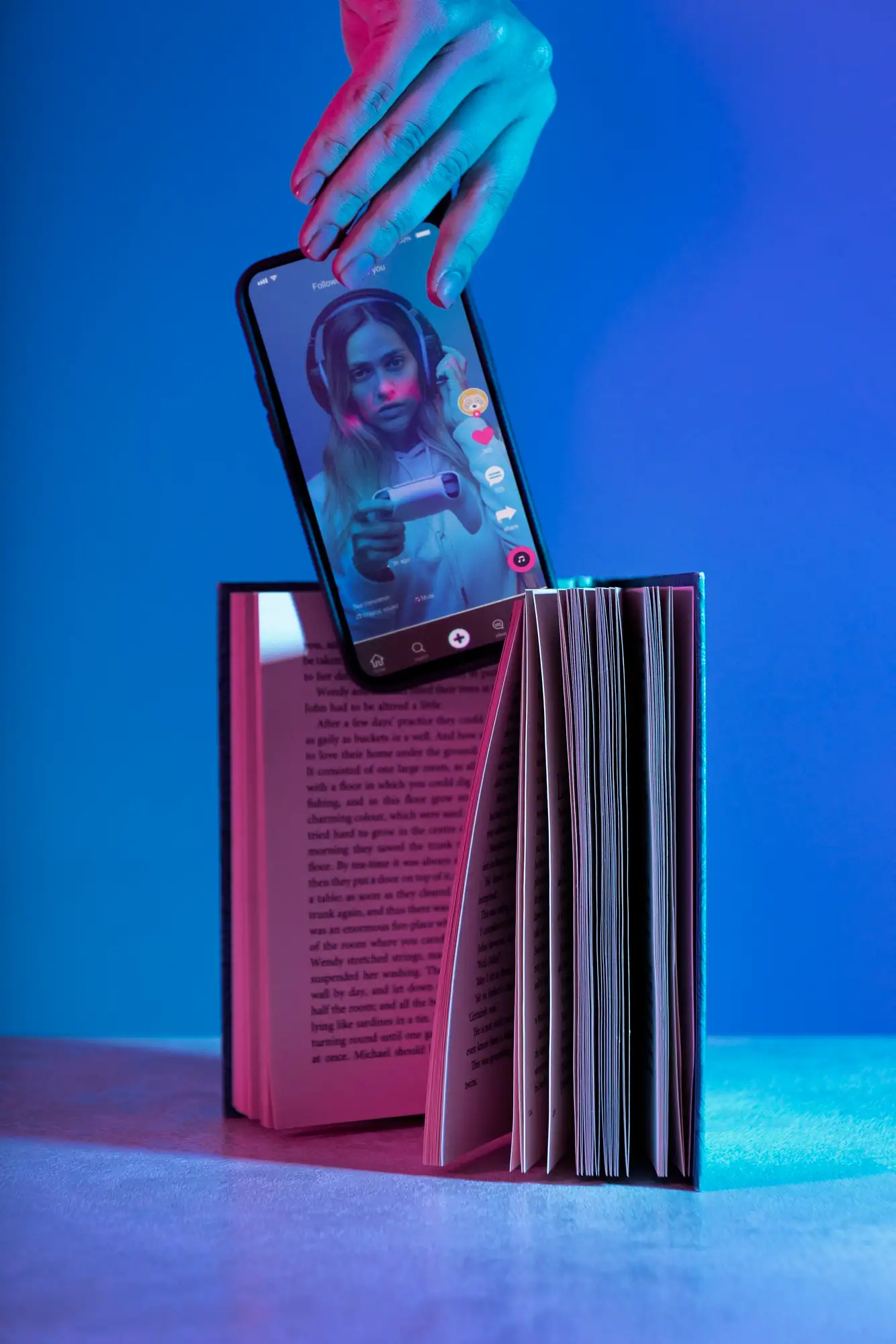 Livre ouvert avec un smartphone ins&eacute;r&eacute; entre les pages, affichant une vid&eacute;o de jeu sur les r&eacute;seaux sociaux, &eacute;clair&eacute; par une lumi&egrave;re n&eacute;on bleut&eacute;e