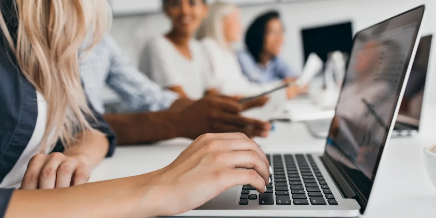 3711 1500X750 | Cekome Gros plan sur une main tapant sur un ordinateur portable, avec un groupe de collègues flous en arrière-plan en réunion
