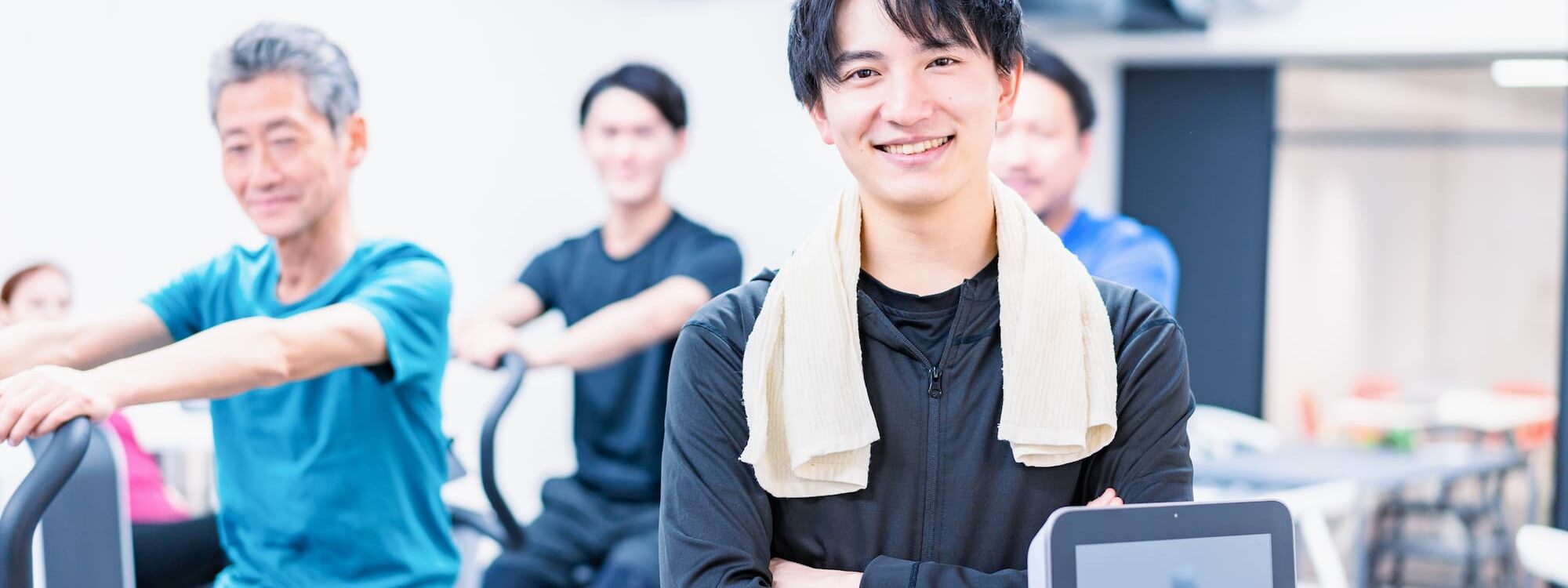COACH SPORTIF SOURIANT DANS UNE SALLE DE SPORT AVEC DES PERSONNES SENTRAINANT EN ARRIERE PLAN 2000X750 | Cekome Coach sportif souriant dans une salle de sport avec des personnes s'entraînant en arrière-plan.