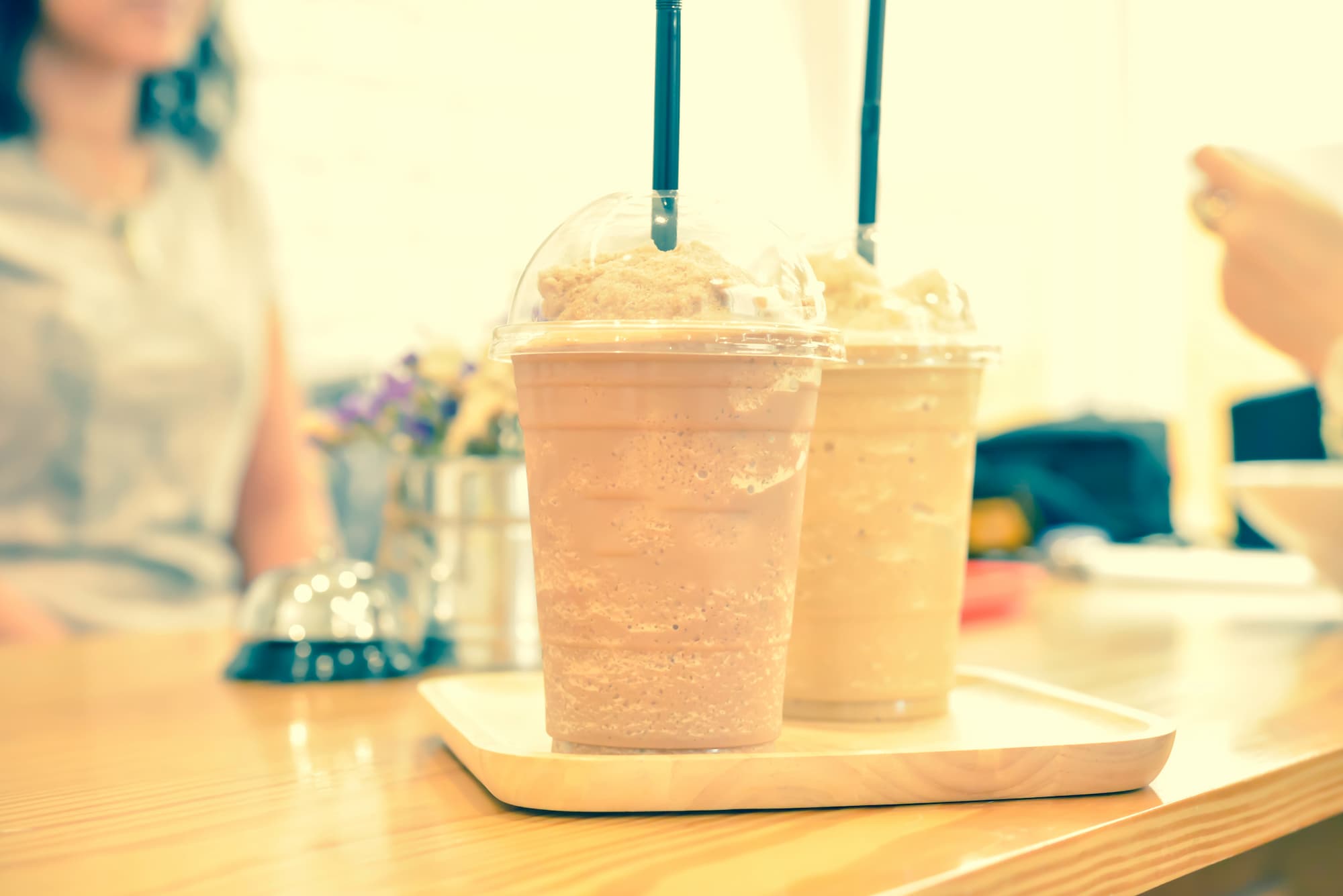 DEUX CAFES FRAPPES DANS DES GOBELETS EN PLASTIQUE AVEC PAILLES POSES SUR UN PLATEAU EN BOIS DANS UN CAFE | Cekome Deux cafés frappés dans des gobelets en plastique avec pailles, posés sur un plateau en bois dans un café