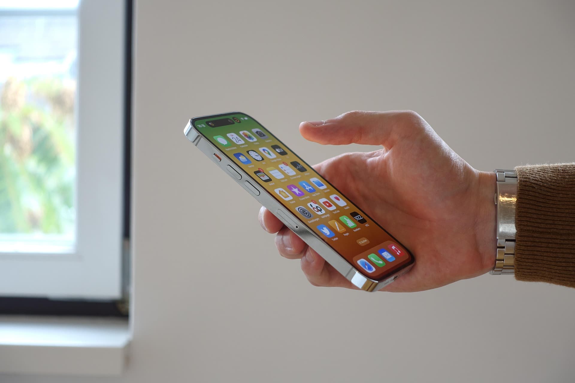 MAIN TENANT UN IPHONE AVEC ECRAN DACCUEIL AFFICHE PRES DUNE FENETRE | Cekome Main tenant un iPhone avec écran d’accueil affiché, près d’une fenêtre