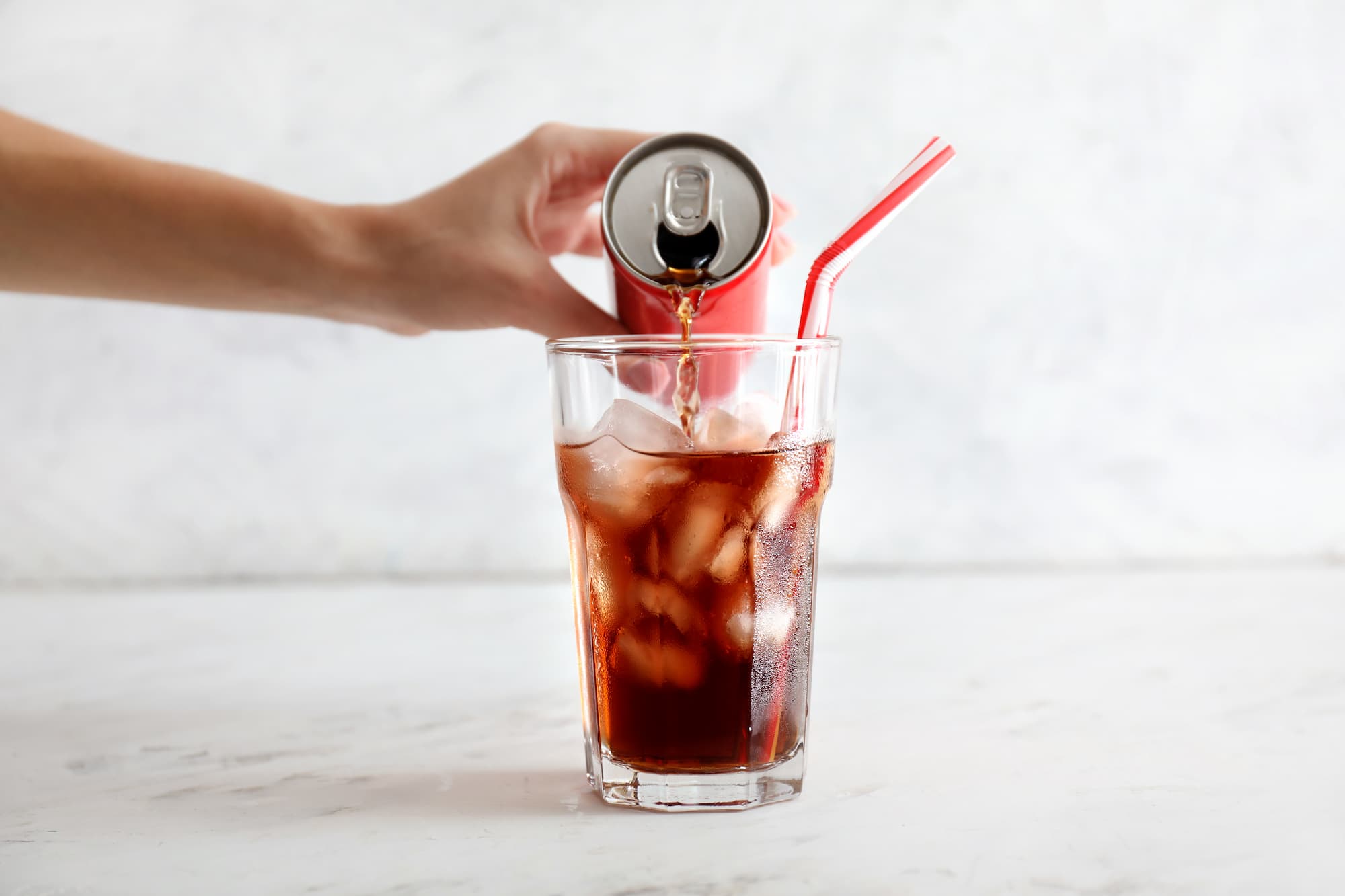 Main versant une boisson gazeuse d'une canette rouge dans un verre avec gla&ccedil;ons et pailles rouges