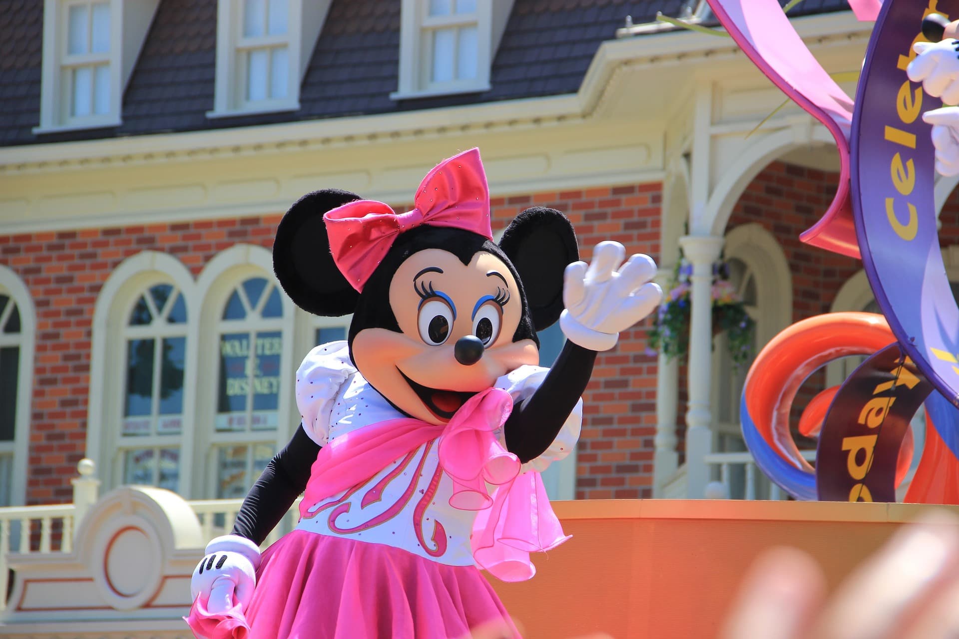 Minnie Mouse en robe rose saluant lors d'une parade Disney devant un b&acirc;timent victorien