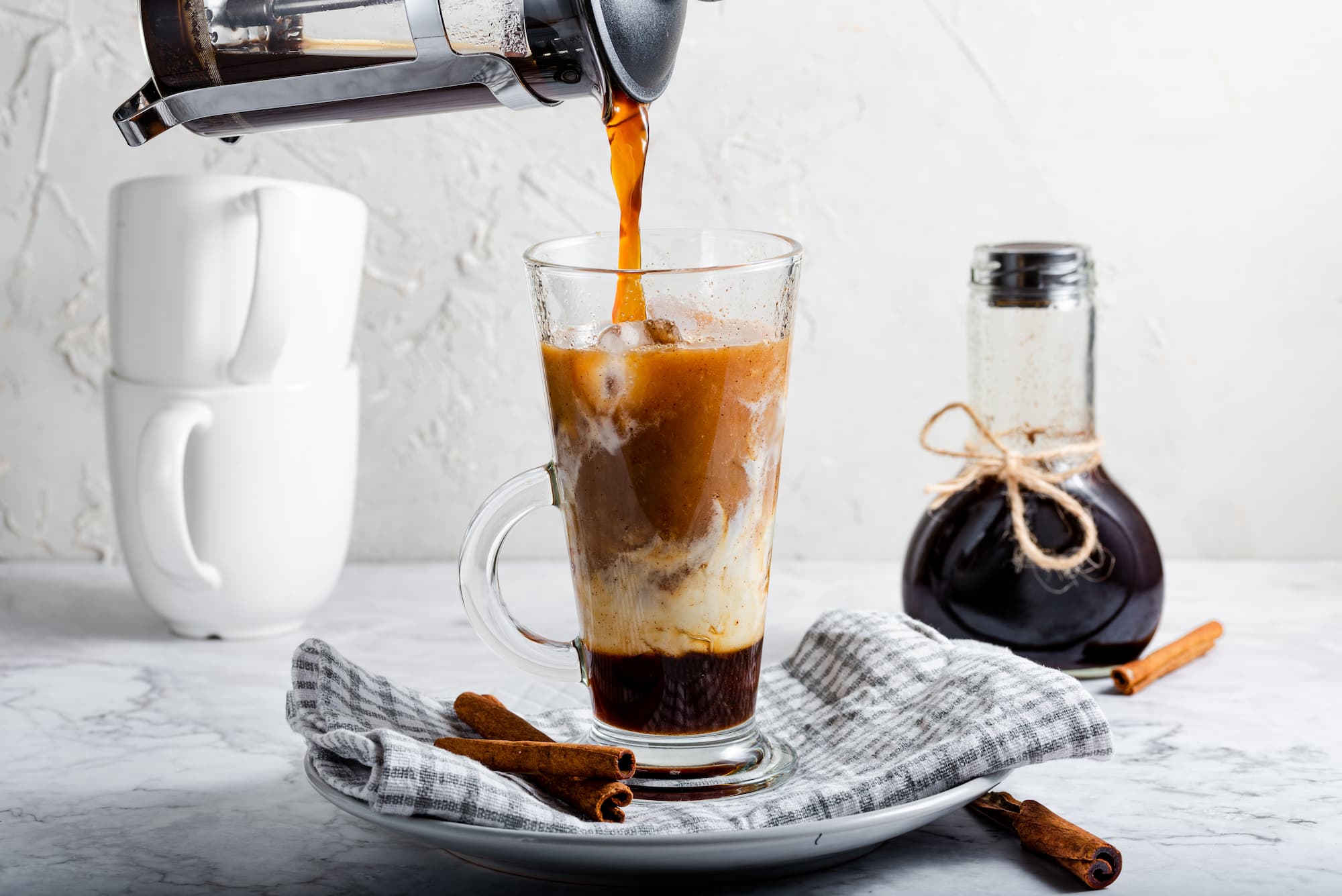VERSEMENT DE CAFE CHAUD DANS UN VERRE TRANSPARENT CONTENANT DU LAIT ET DE LA CREME ENTOURE DE BATONS DE CANNELLE | Cekome Versement de café chaud dans un verre transparent contenant du lait et de la crème, entouré de bâtons de cannelle