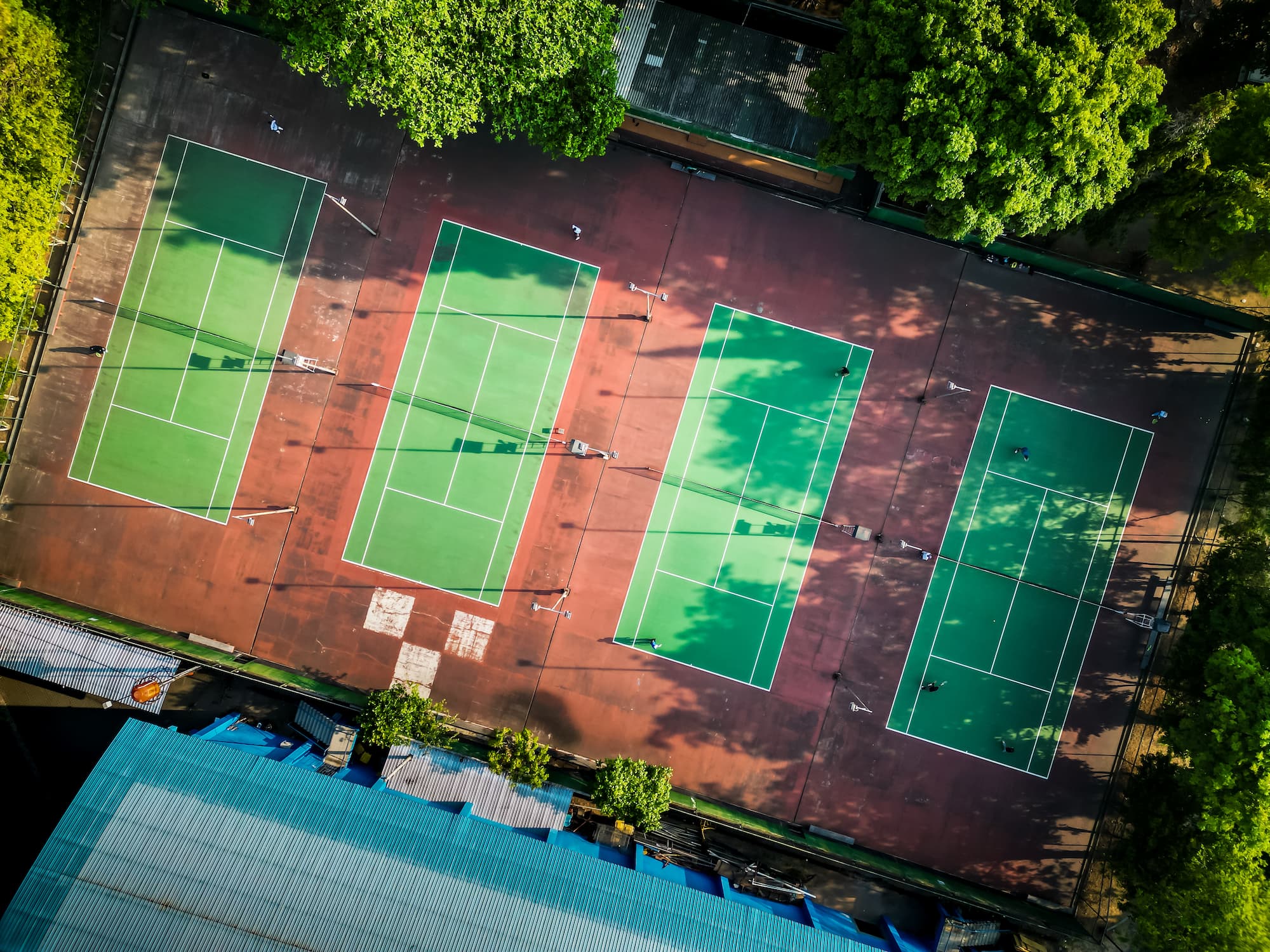 VUE AERIENNE DE PLUSIEURS TERRAINS DE TENNIS EN EXTERIEUR ENTOURES DARBRES | Cekome Vue aérienne de plusieurs terrains de tennis en extérieur entourés d’arbres.