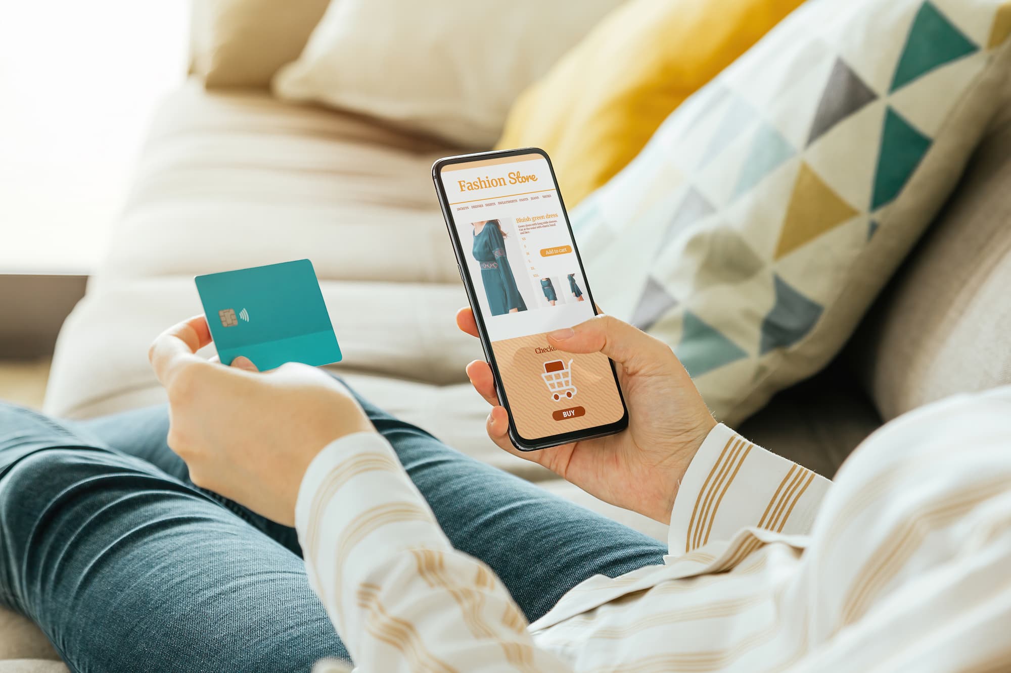 PERSONNE ALLONGEE SUR UN CANAPE FAISANT DU SHOPPING EN LIGNE VIA UN SMARTPHONE TENANT UNE CARTE BANCAIRE TURQUOISE | Cekome Personne allongée sur un canapé faisant du shopping en ligne via un smartphone, tenant une carte bancaire turquoise.