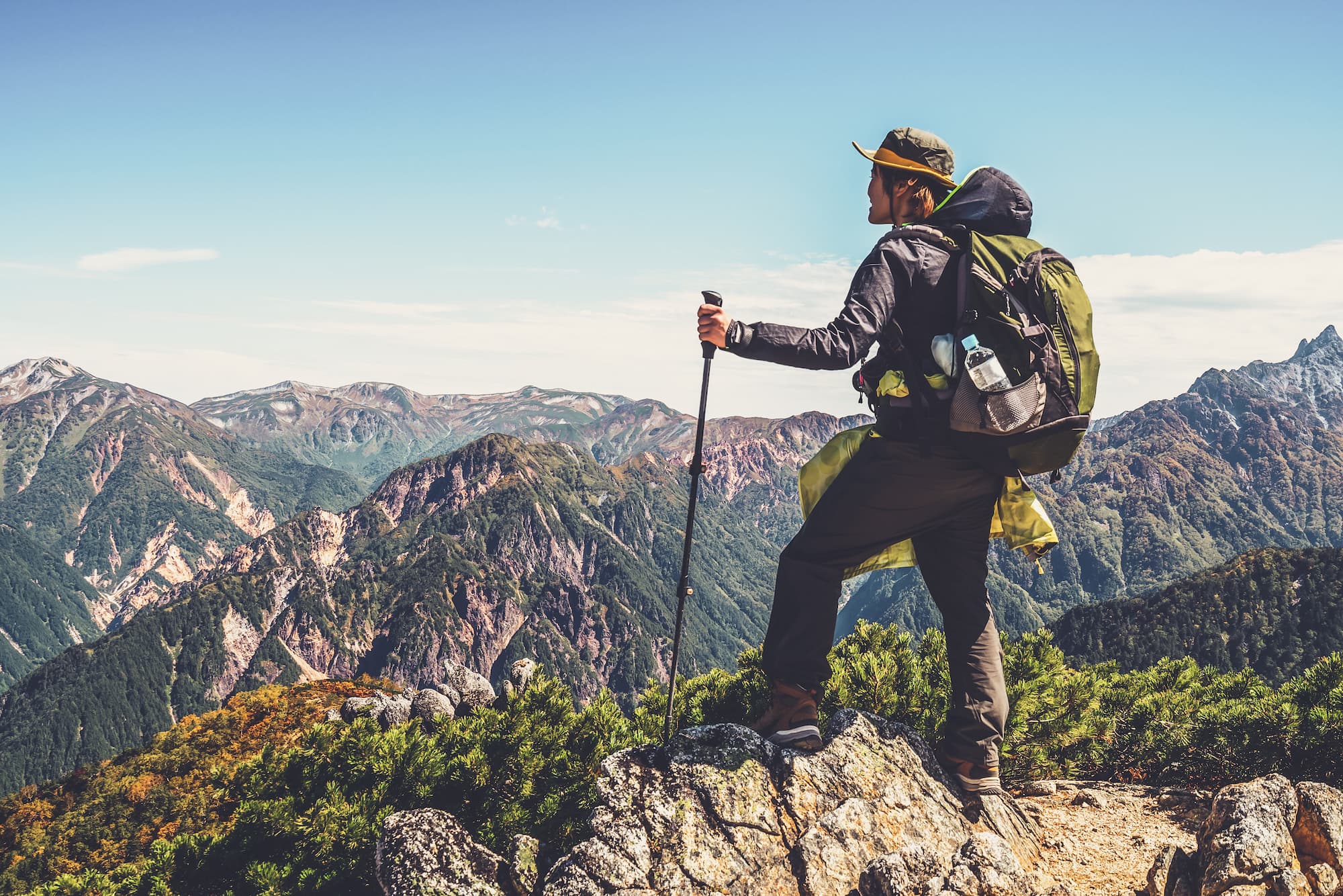 ADOBESTOCK_264777661 | Cekome Randonneur au sommet d'une montagne admirant un paysage de crêtes verdoyantes sous un ciel bleu clair.